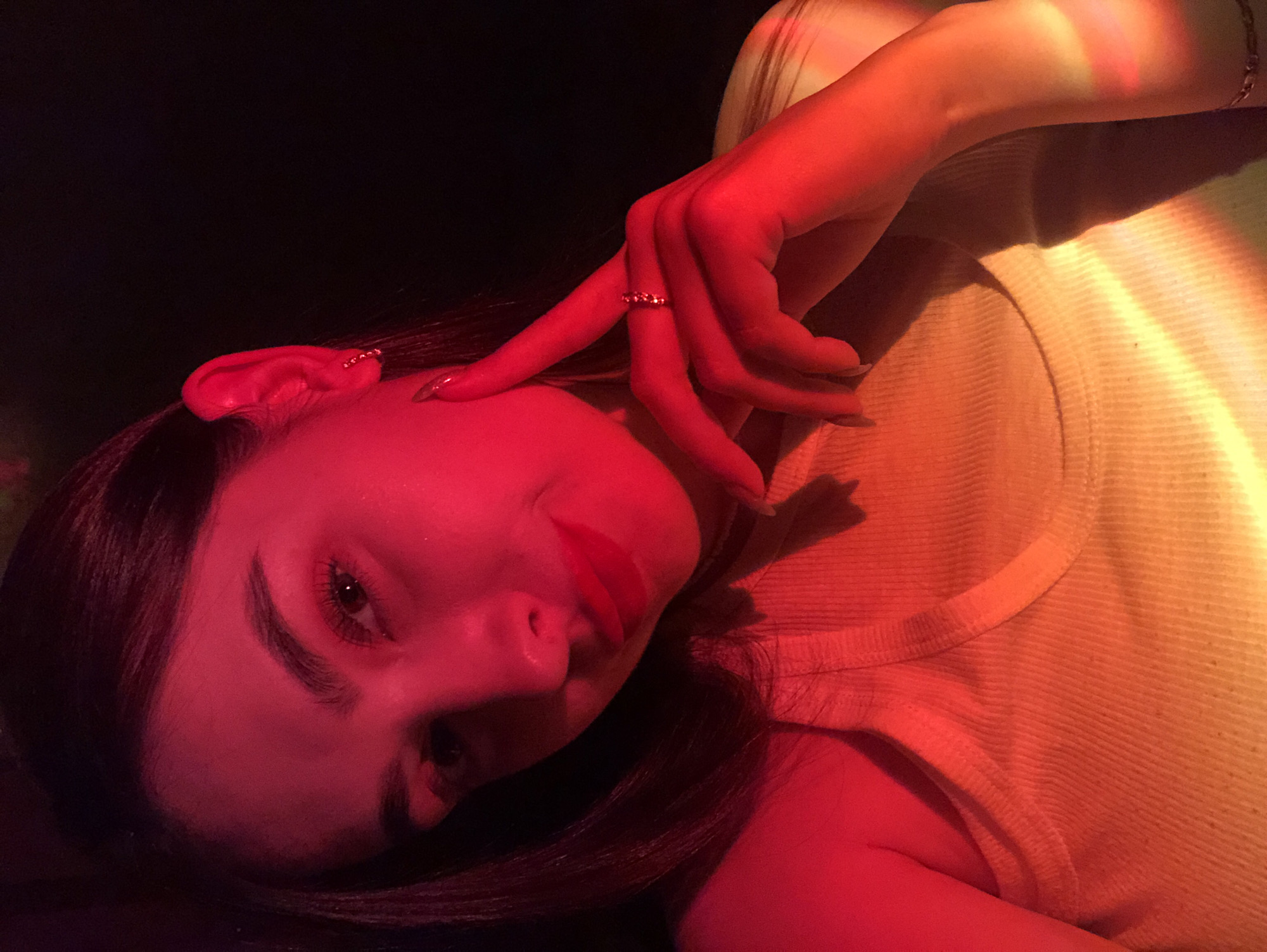 A person with light skin, dark hair, and full lips is lying down, head tilted towards the left. A hand is raised near the face, with a ring visible on one finger. The lighting is predominantly red and warm, with a hint of yellow on the side. The person appears to be wearing a light-colored ribbed tank top. The background is dark and mostly obscured. The setting is Noviy, Russia.