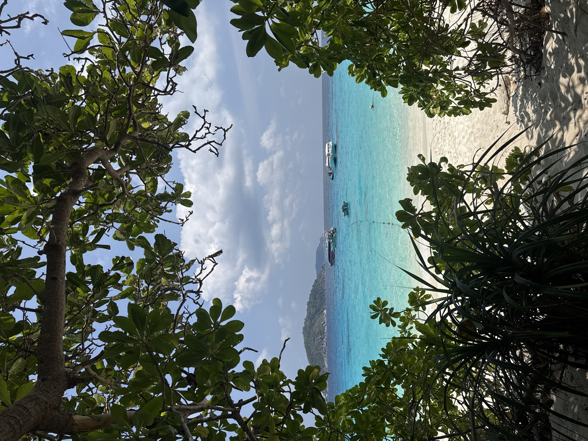 The image depicts a beach scene in Lam Kaen, Thailand. The view is framed by foliage in the foreground. Turquoise water stretches toward the horizon where clouds partially obscure the sky. Several boats are visible in the water. A sandy beach is present to the right of the water. An island can be seen in the distance.