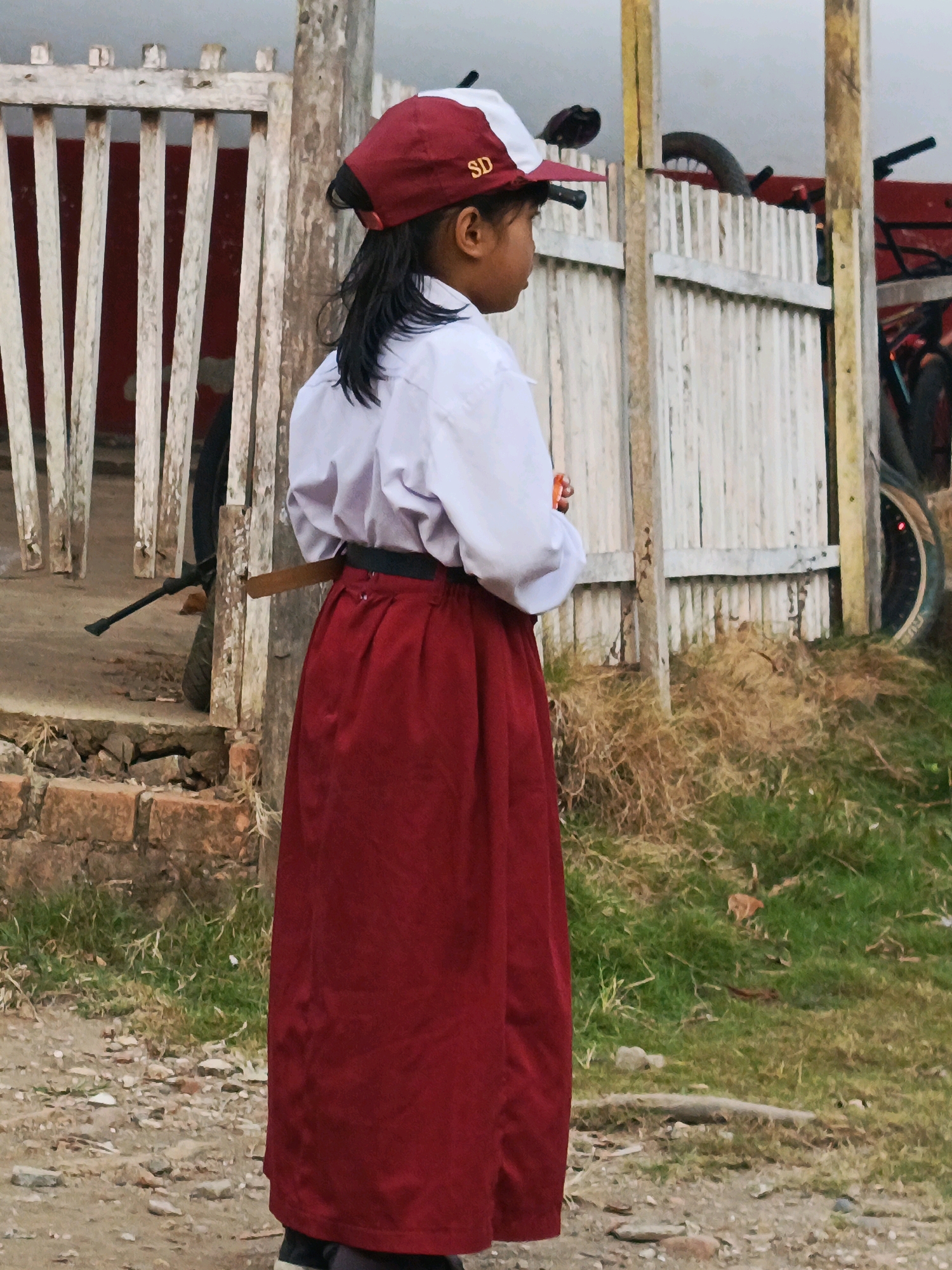 The image shows a young girl standing on a dirt path in front of a rustic wooden fence. She is wearing a white collared shirt, a dark belt, and a long, flowing red skirt. On her head, she wears a red and white baseball cap with "SD" embroidered on the front. Her dark hair is pulled back. To the left of the girl, a dark bicycle wheel is visible behind the fence. Behind the girl, a red and white structure with more wooden fencing is present. The ground is covered with gravel and sparse green and dry grass. In the background, a larger red object, possibly a vehicle or building, is partially visible. The overall impression is a candid shot of a child in an outdoor, rural or semi-rural setting.