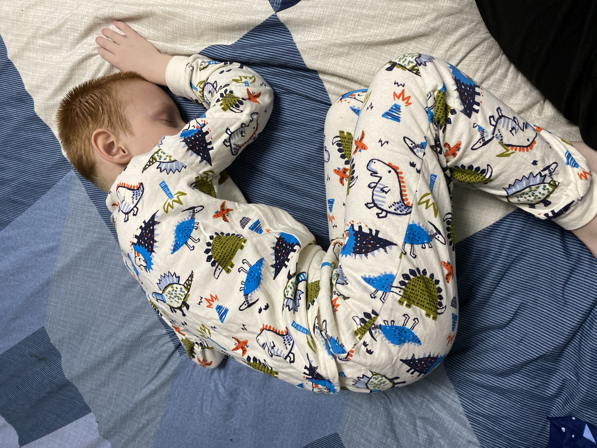 A young child with red hair is sleeping on a bed covered with a blue and white patterned quilt. The child is wearing white pajamas with a dinosaur pattern. The location is not identifiable from the image.