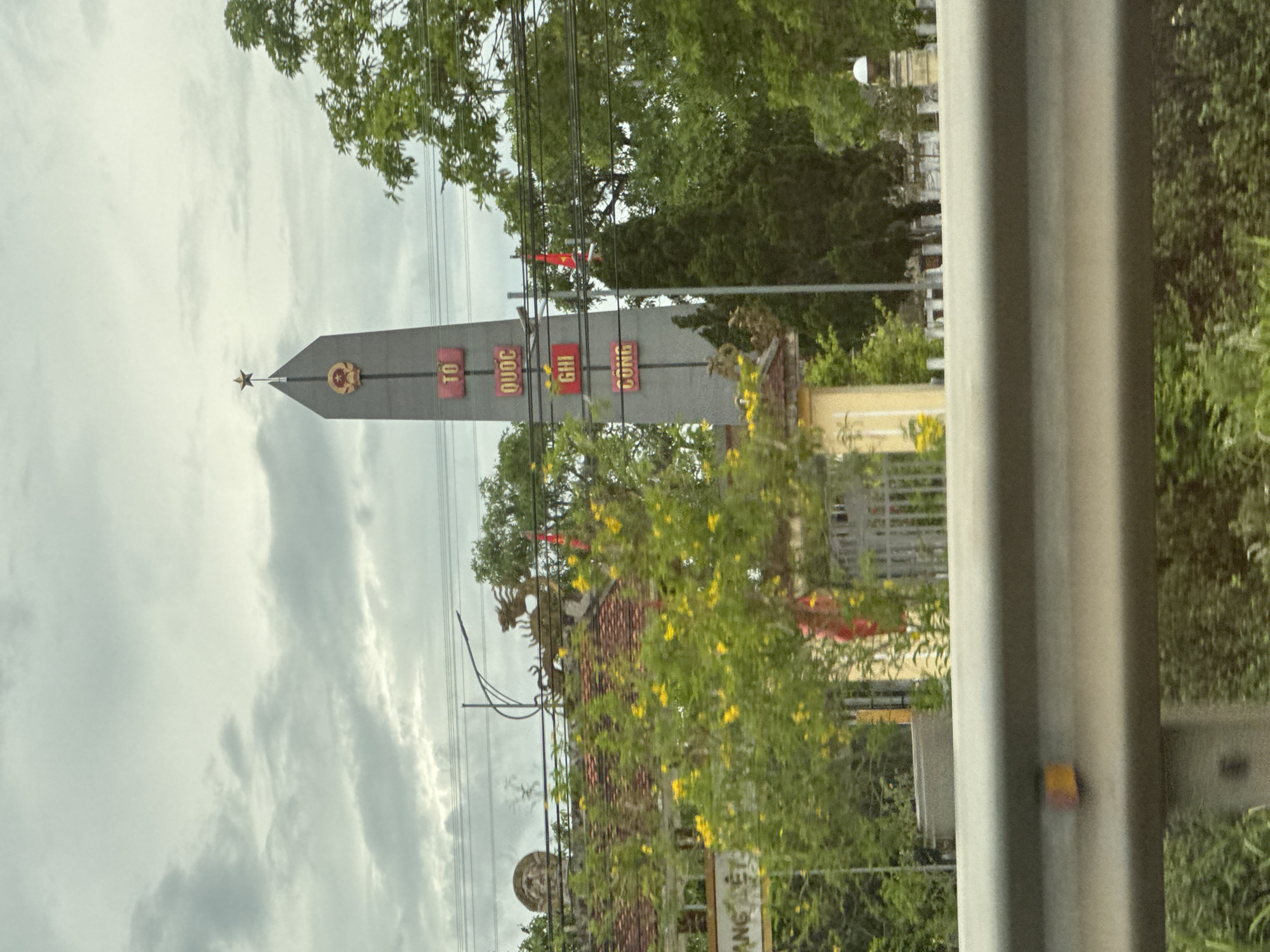 The image shows a monument in Hải Lăng Commune, Vietnam. The monument is a tall, gray obelisk topped with a star. The obelisk has inscriptions in Vietnamese that read "TỔ QUỐC GHI CÔNG" which translates to "The Nation Remembers Your Merits." A red flag with a yellow star, the flag of Vietnam, flies behind the monument. The monument is surrounded by trees and a cloudy sky. In the foreground, a portion of a metal guardrail is visible. The overall scene suggests a solemn memorial site.