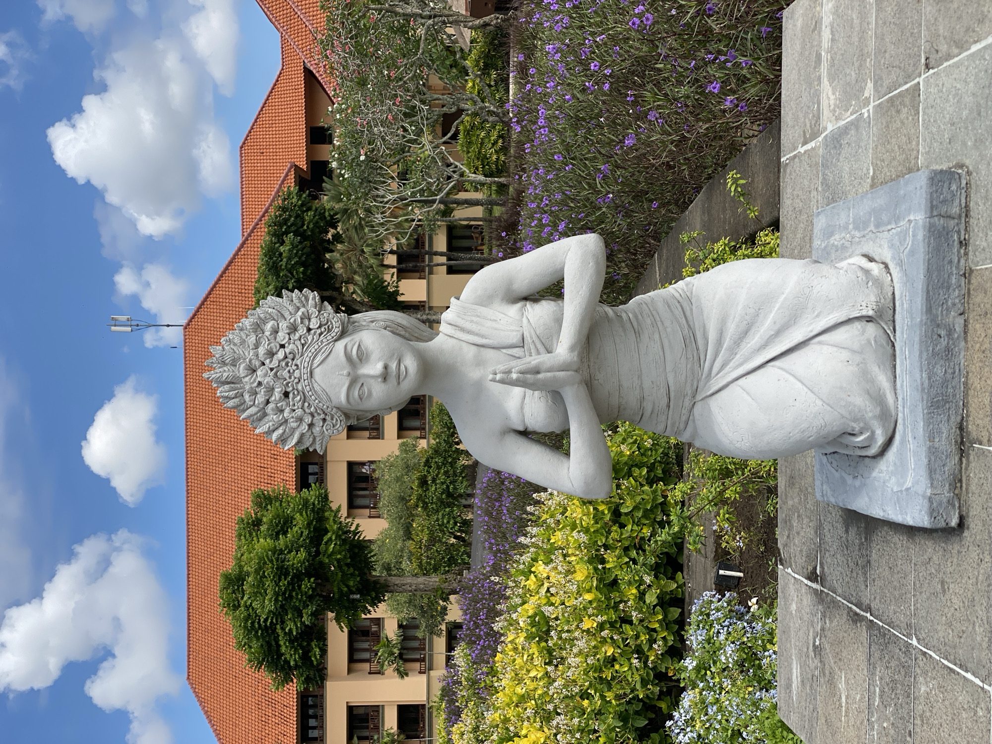 A gray, concrete statue of a kneeling woman with her hands pressed together in a traditional greeting pose stands centered in the foreground. The statue wears an ornate headdress and a draped garment. The statue is positioned on a paved stone area in front of a lush garden filled with yellow and purple flowers. Behind the garden, a multi-story building with orange tiled roofs and light-colored walls is visible. Trees and other foliage surround the building. The sky above is blue with scattered white clouds. The scene appears to be in a resort or hotel setting in Kuta Selatan, Indonesia, a popular tourist area known for its beaches and resorts.