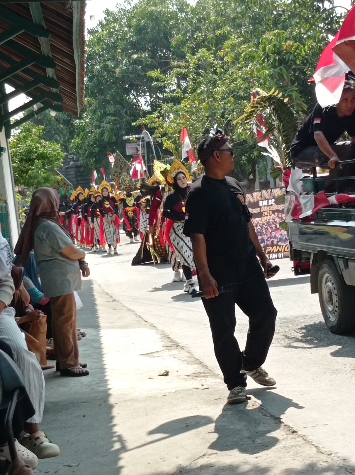 In Maribaya, Indonesia, a group of people are participating in a parade. The foreground shows a man in a black t-shirt and black pants walking alongside a flatbed truck. He is holding a microphone in his right hand. Behind him, a procession of individuals in traditional costumes with elaborate headwear and red and white accents are marching down a paved road. Several red and white Indonesian flags are visible throughout the scene, adorning the truck and being carried by some participants. The parade is taking place outdoors, with lush green trees and a building with a wooden roof visible in the background. The sun is shining, casting shadows on the ground. A sign on the back of the truck displays "KARNAVAL RW.02" and other text.