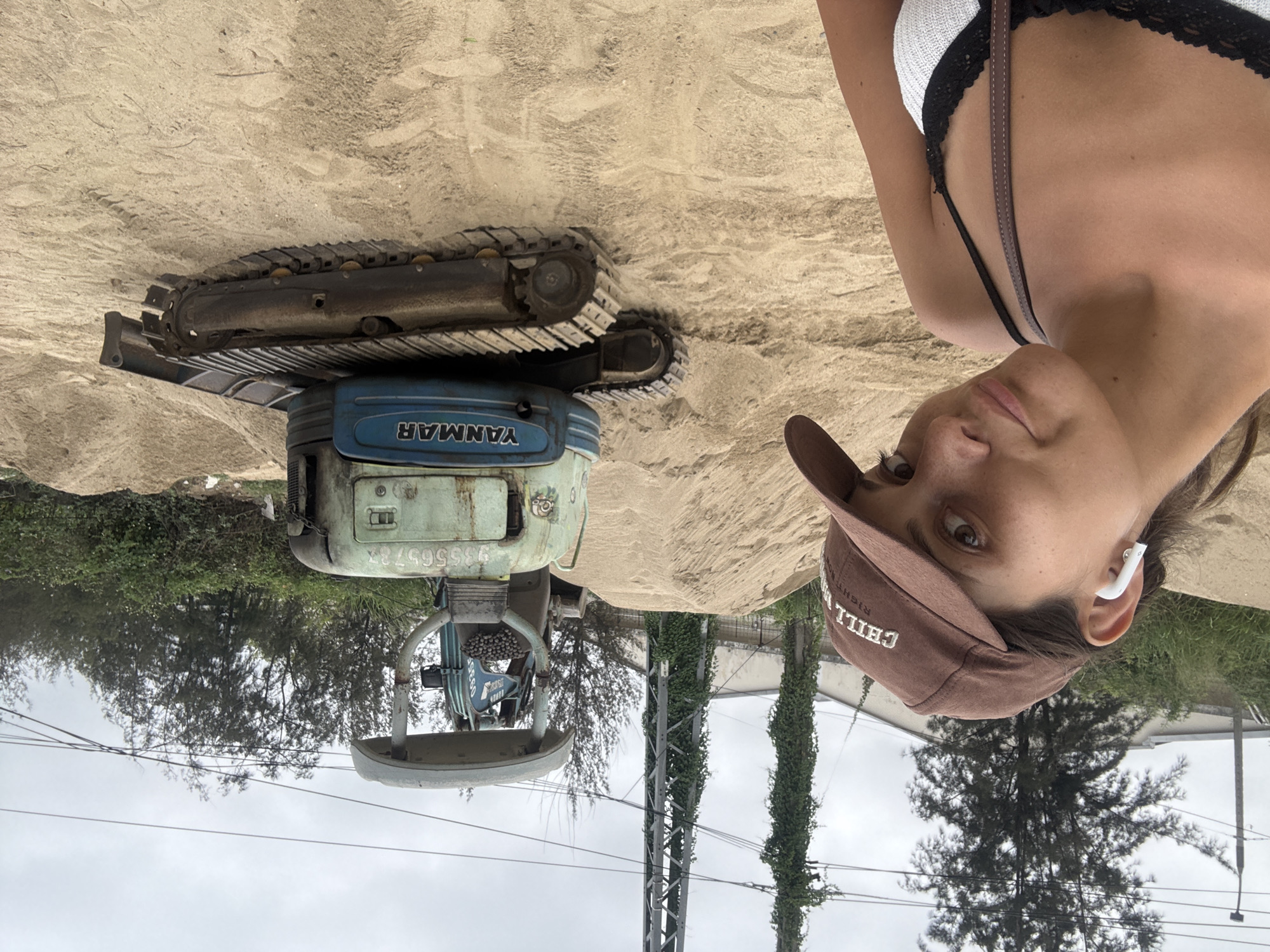 A rotated outdoor photograph depicts a woman posing in front of a small, used Yanmar excavator on a sandy surface in Đà Nẵng, Vietnam.
The excavator, primarily blue and grey, shows signs of wear and rust. The woman is wearing a brown baseball cap, a white and black top, and an earbud in her right ear.
Background details include trees, utility poles covered in climbing plants, and a cloudy sky. The environment suggests an urban or suburban area undergoing construction or landscaping.