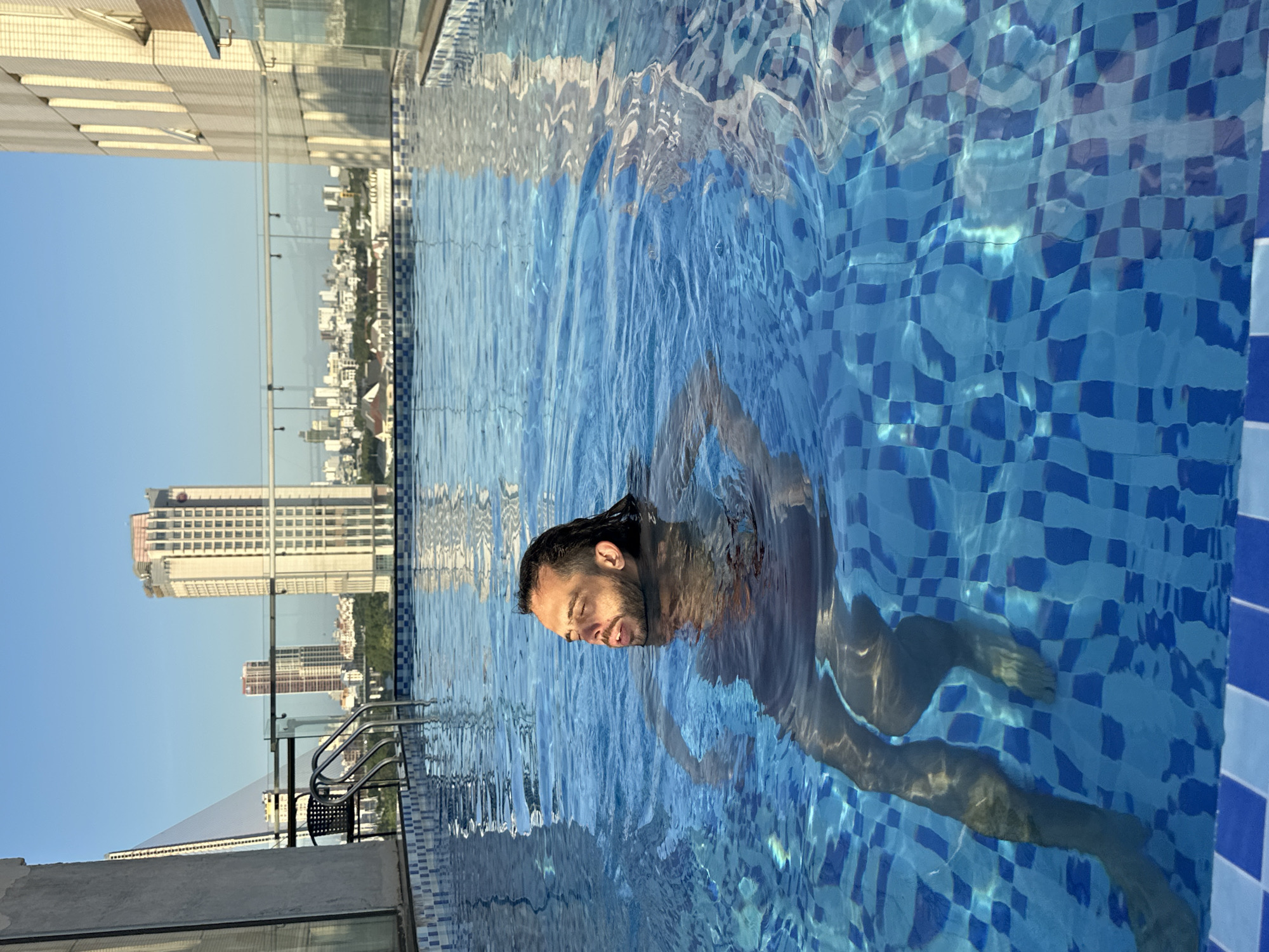 A man with dark hair and a beard swims in a rooftop swimming pool in Da Nang, Vietnam. The water is clear blue with a checkered tile bottom. The man's head and shoulders are visible above the water, and his body is submerged. In the background, the cityscape of Da Nang is visible, with tall buildings and a bridge. The sky is clear and blue. The scene is illuminated by natural sunlight.