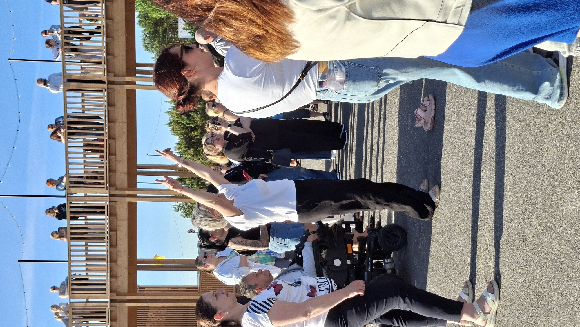 A crowd of people stands on an asphalt surface near a wooden structure. A woman in a white shirt and black pants stands in the foreground, with her arms raised and fingers pointed upwards. To her left, a man in a wheelchair is being attended to by a woman in a white and black striped shirt. Behind them, more people are visible, some standing on an elevated wooden walkway. The scene is outdoors, with a clear blue sky and trees in the background. The wooden structure appears to be a part of a public space, possibly a park or an event venue. The presence of a wheelchair suggests accessibility features are present. The overall atmosphere appears to be social and possibly celebratory, given the raised arms and the gathering of people. The location in Tallinn, Estonia is not explicitly identifiable from the visual information alone.