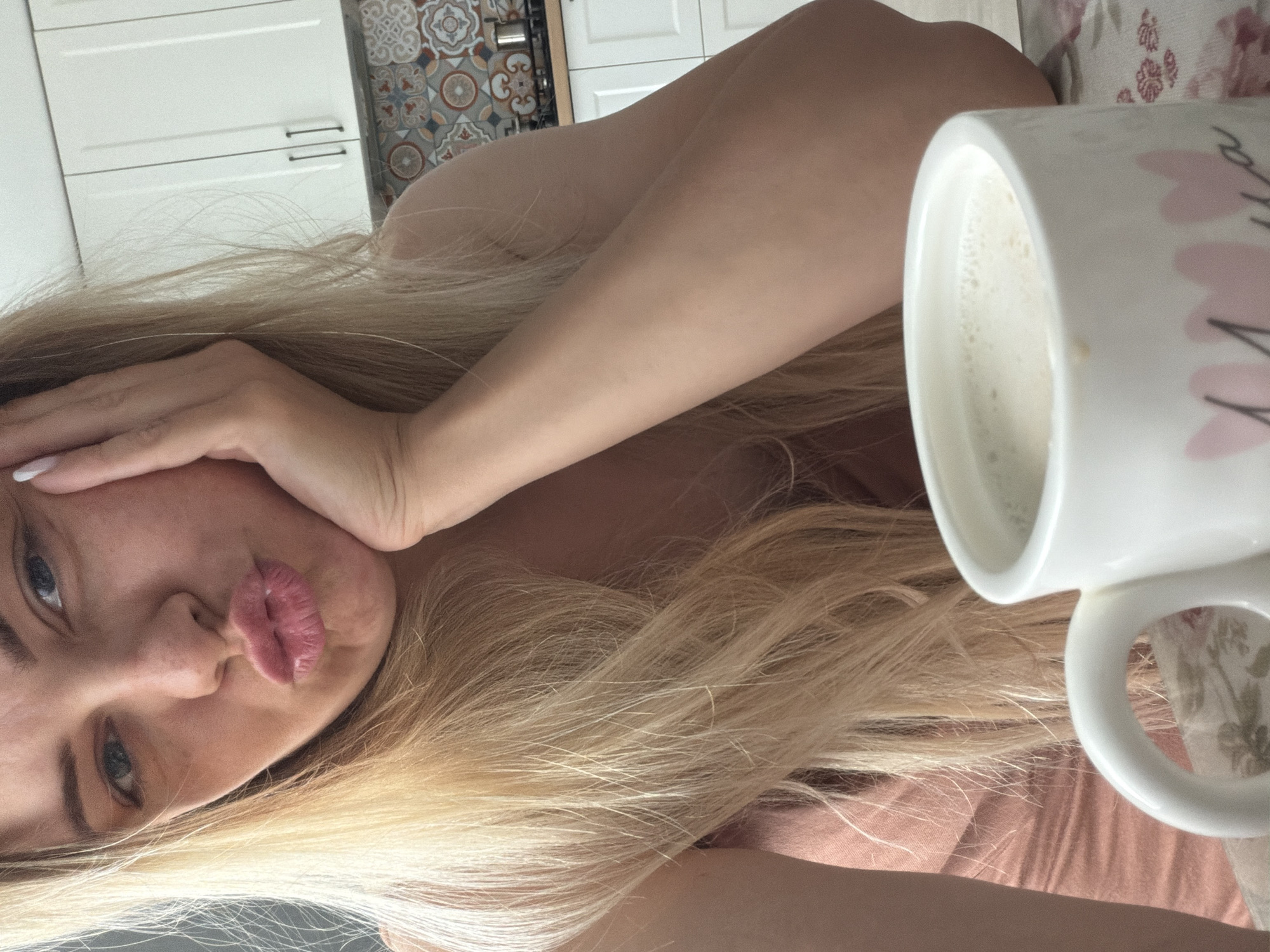 A woman with blond hair and blue eyes is shown in a close-up shot, tilting her head slightly and making a kissing face. Her hand is near her face, with white nail polish visible on her nails. A white mug filled with a light-colored liquid is positioned in the foreground. The background includes white kitchen cabinets and a patterned tile backsplash.
It is not possible to determine if the photo was taken in Votkinsk, Russia, as no specific location details are visible.