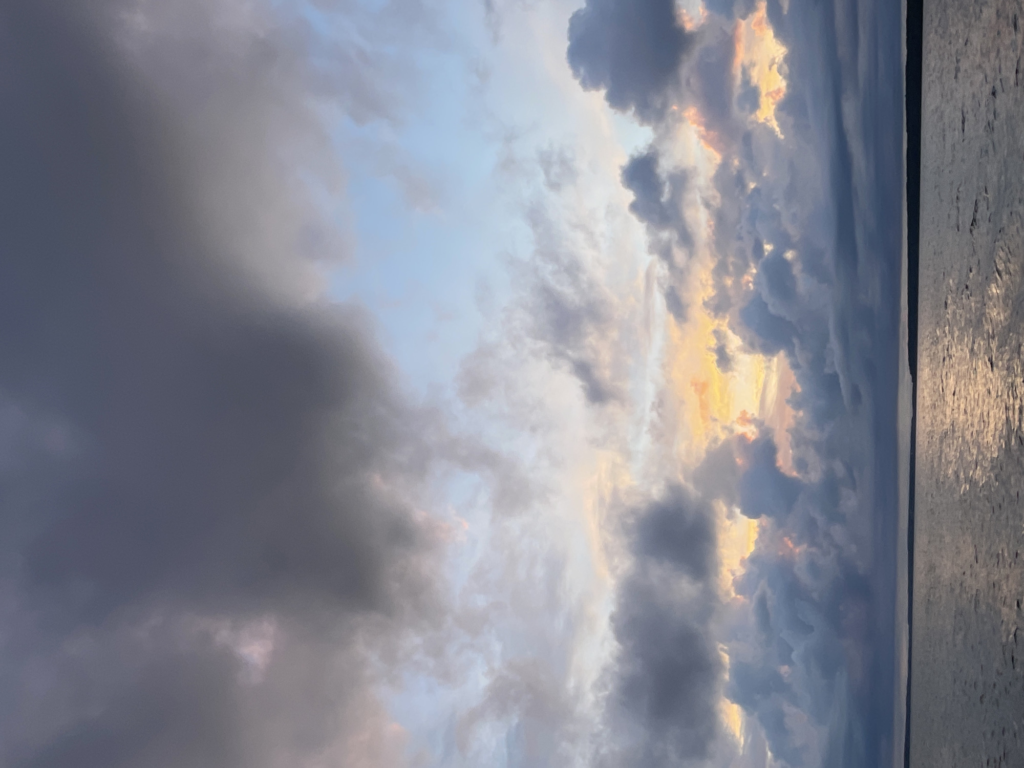 The image depicts a body of water under a dramatic sky filled with dark, heavy clouds. Streaks of pale yellow and pink light are visible through gaps in the clouds, suggesting either sunrise or sunset. The water is choppy, reflecting the colors of the sky. A dark, low-lying landmass is visible in the distance, creating a horizontal line against the horizon. The scene appears to be a natural landscape.