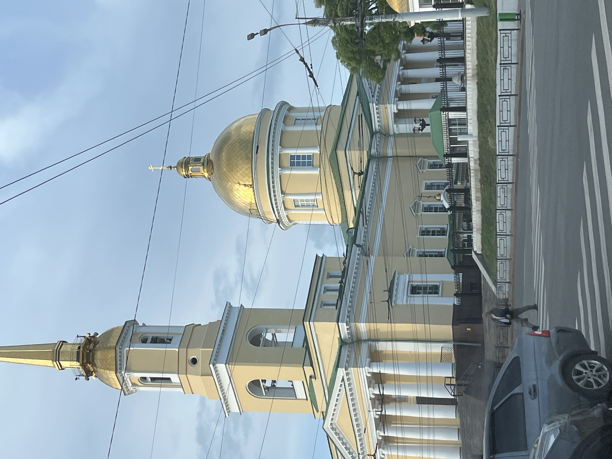 The image shows St. Michael's Cathedral in Izhevsk, Russia, from a low angle, partially obstructed by a grey car in the foreground. A pedestrian is crossing a street near the cathedral. The cathedral features a tall bell tower and a central dome, both topped with gold. Overhead electrical lines are visible against the sky.