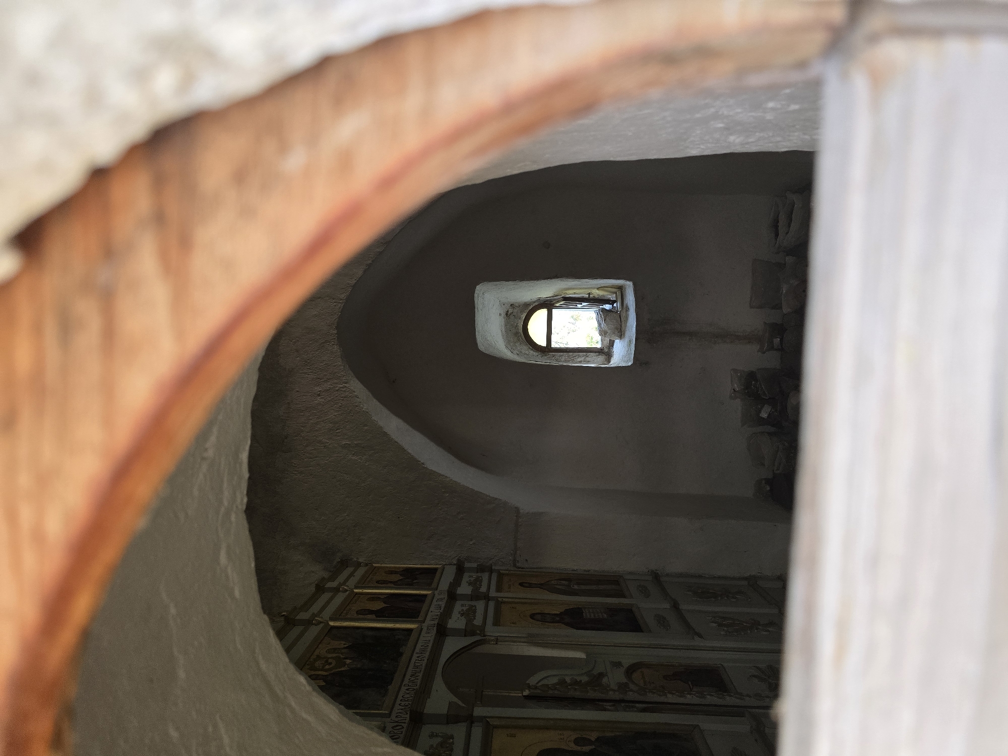The image depicts the interior of what appears to be a church or chapel, possibly located in Medun, Montenegro, due to the presence of religious iconography. The scene is viewed through an arched opening with wooden trim.

In the foreground, out of focus, is a light-colored horizontal surface, possibly a bench or ledge. Beyond this, the interior features vaulted, rough-textured walls. To the left, a wall is adorned with religious icons, likely part of an iconostasis. These icons are arranged in multiple rows and columns, depicting figures in traditional religious attire.

Further into the interior, a small, arched window is visible in the back wall. The window allows natural light to illuminate the space, and through it, greenery and possibly sky can be seen. The area below the window appears to have some scattered stones and possibly bags or fabric bundles. The overall atmosphere is one of aged architecture and religious significance.