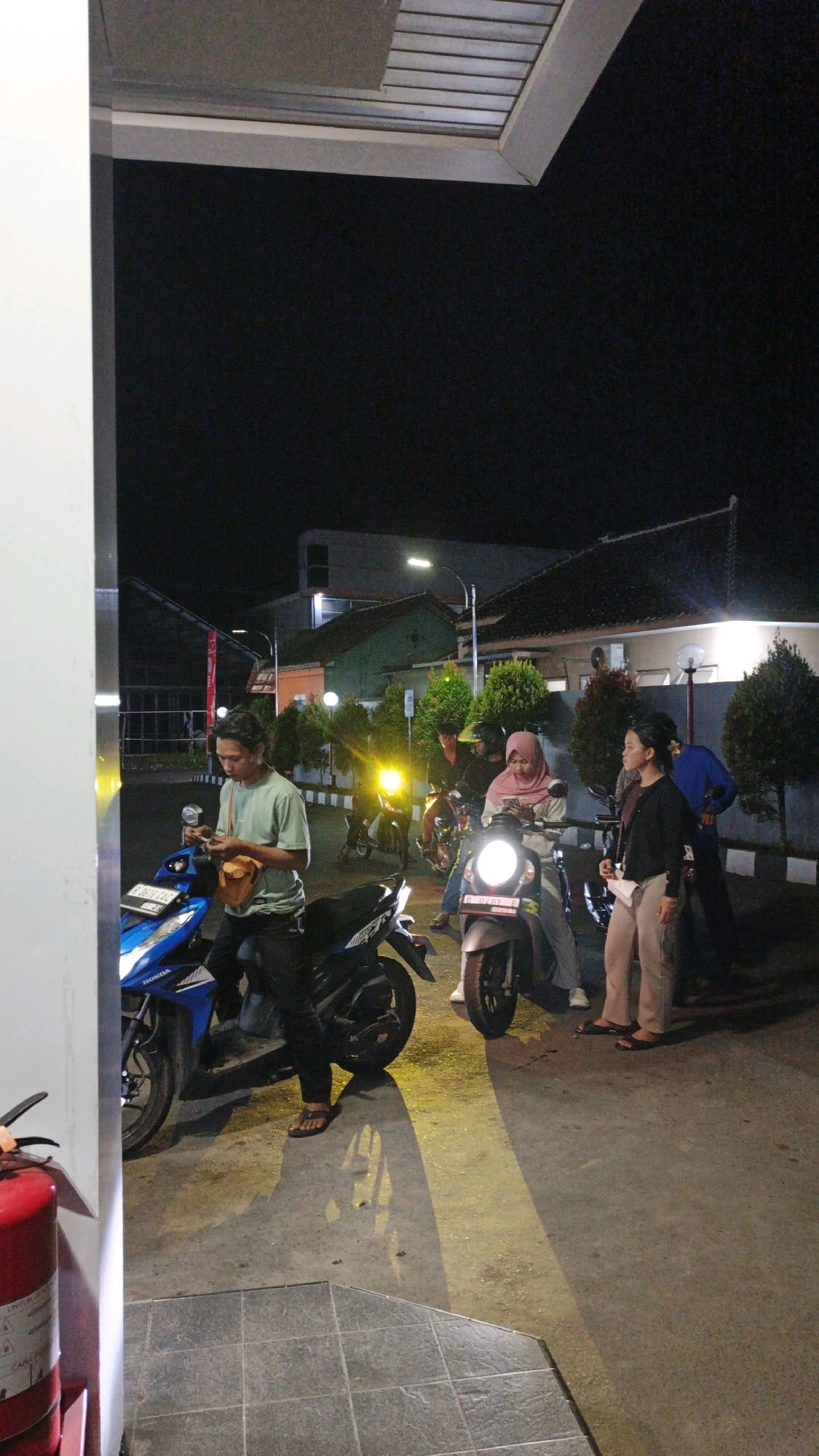 In Karangjaladri, Indonesia, at night, a group of people are gathered around motorcycles under an overhang, possibly a gas station or covered parking area. The foreground features a bright blue and black motorcycle, with a person in a light green shirt and dark pants sitting on it, holding a smartphone and a small brown bag. To the right, another motorcycle has a person in a pink hijab and grey pants sitting on it, also looking at their phone. Standing next to them is a person wearing a black shirt and light brown pants. Further back, other individuals and motorcycles are visible, with bright headlights illuminating the scene. The environment appears to be an outdoor paved area with some greenery and buildings in the background. A red fire extinguisher is visible on the left edge of the image.