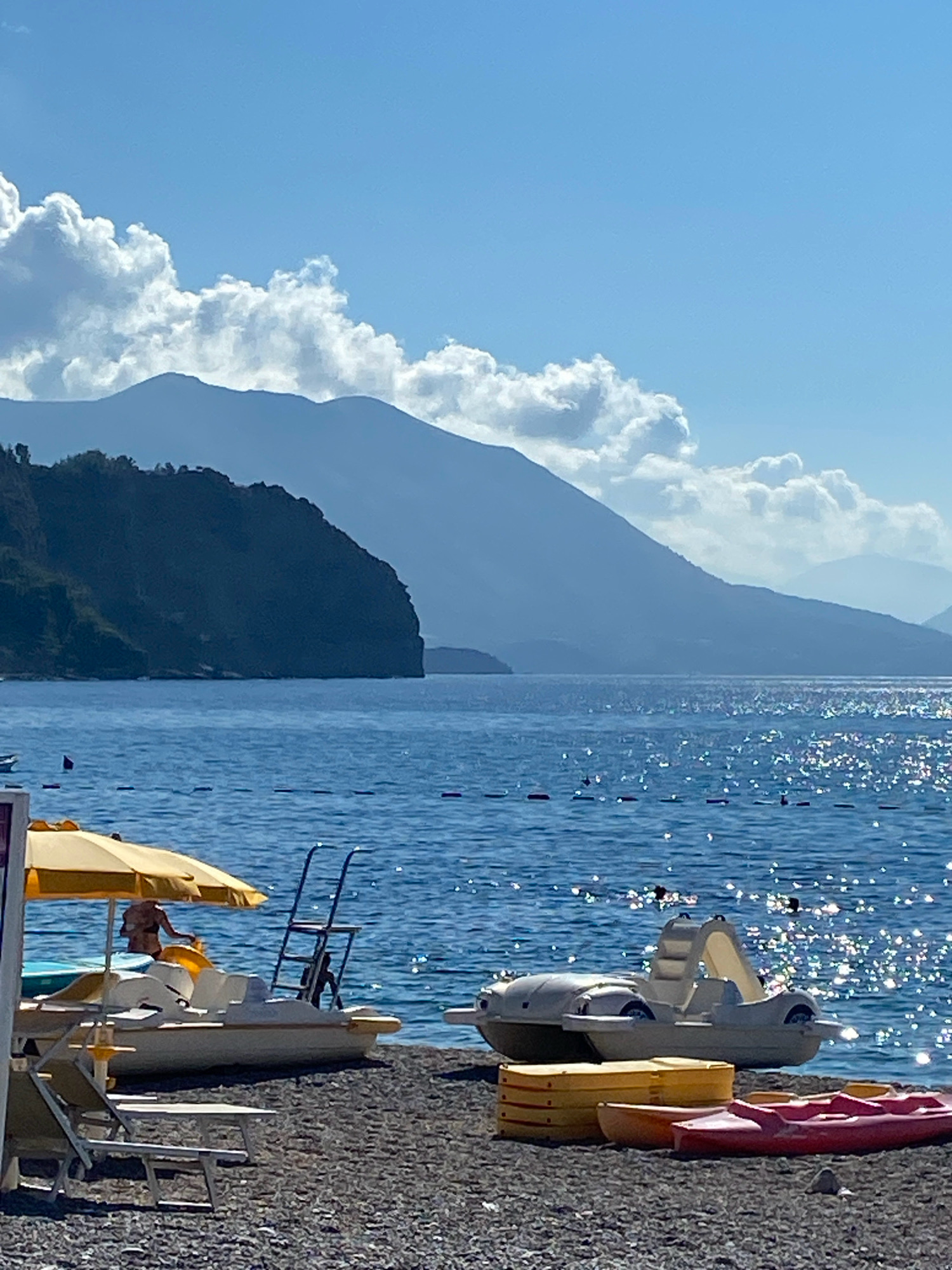 The image depicts a sun-drenched beach scene at the sea, with mountains in the background. In the foreground, a pebble beach is lined with colorful kayaks and pedal boats, suggesting recreational activities. A yellow beach umbrella is set up, providing shade. To the left, two lounge chairs are positioned on the beach. The water is a deep blue, reflecting the sunlight with a shimmering effect. In the distance, a dark, tree-covered peninsula juts out into the sea, and behind it, layers of hazy blue mountains rise against a bright blue sky dotted with fluffy white clouds. The overall impression is one of a peaceful and idyllic summer day at the coast. The location is stated to be in Prijevor, Montenegro.