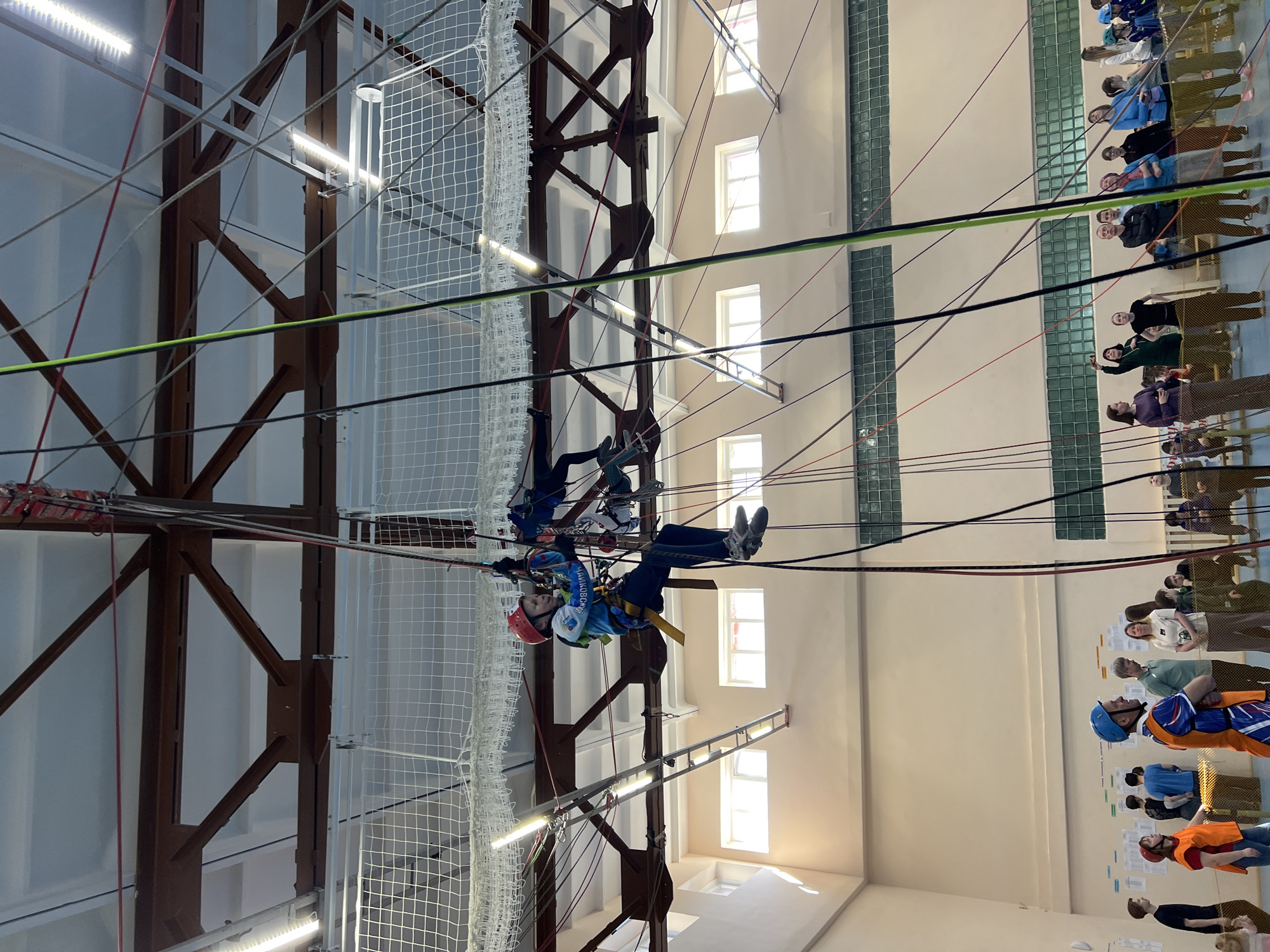 The image shows an indoor climbing structure in Berezniki, Russia. Two individuals are navigating a high-rope course. The person closer to the viewer wears a red helmet, a blue jacket with white lettering, and a yellow harness. The other person wears a blue helmet. They are suspended by ropes and cables connected to an overhead metal framework. A safety net surrounds the structure. In the background, a group of people are observing. The building's interior is lit by rows of fluorescent lights, with windows visible on the right wall.