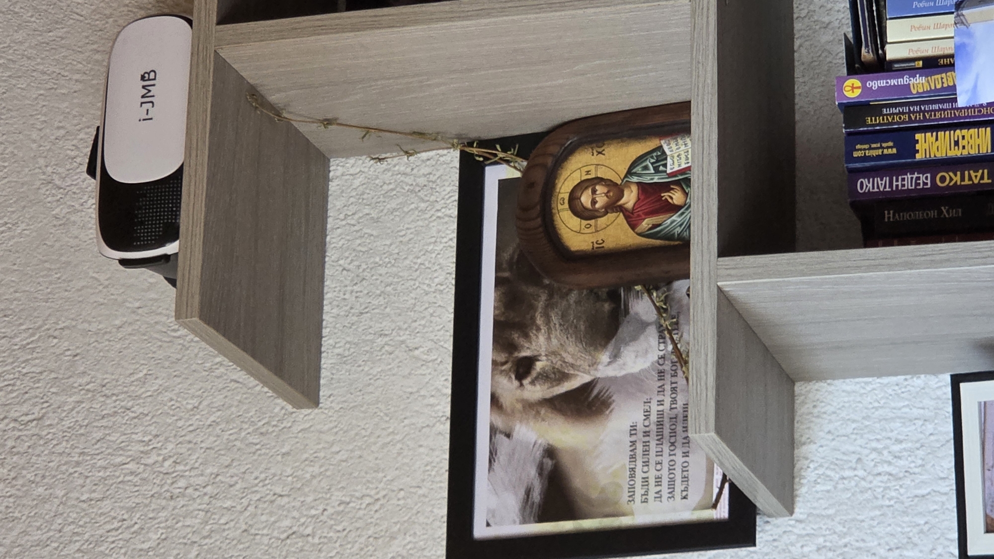 The image shows a close-up of a white textured wall with a light-colored wooden shelf unit against it. On the top shelf, a white and black virtual reality headset with the text "i-JMB" is placed. Below this, on a lower shelf, a framed picture featuring a close-up of a lion's face is visible. To the right of the framed picture, a small wooden-framed icon depicting Jesus Christ is placed. Further down on the shelf, several books are stacked, with spines visible. The books have various titles, some in Cyrillic script. The overall environment suggests an indoor setting, likely a home or office.