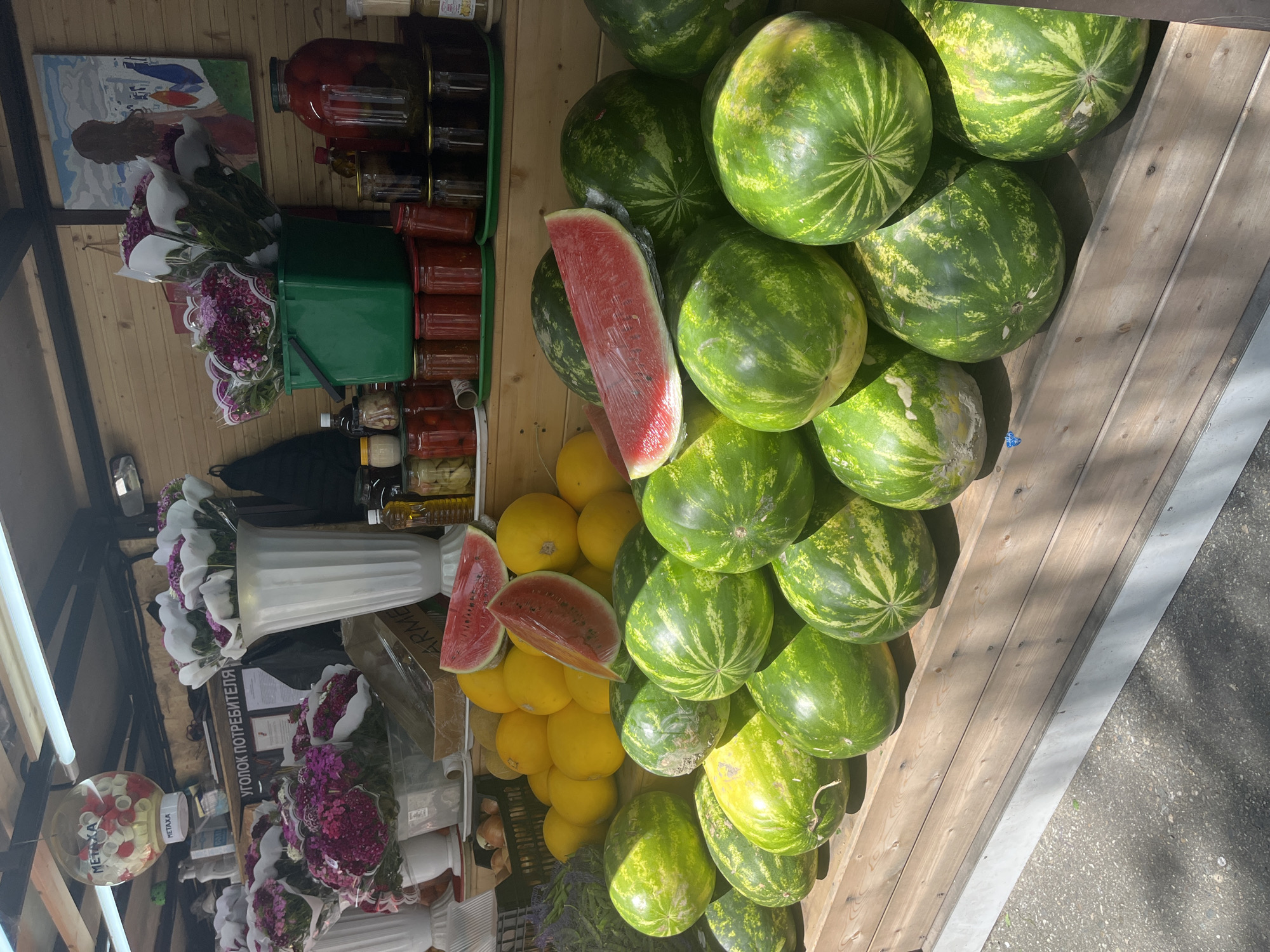 The image captures a vibrant market stall in Sochi, Russia, bustling with fresh produce and various goods under bright daylight. The main focus is a large, abundant display of watermelons, piled high on a wooden stand to the right. These watermelons are green with prominent darker green stripes, and some exhibit lighter yellowish patches. Slices of a ripe red watermelon, wrapped in plastic, are visible among them, hinting at the fruit's freshness.
Below and to the left of the watermelons, numerous round, yellow melons (likely cantaloupes or honeydews) are also stacked. The stall also offers a variety of jarred preserved goods, including what appear to be pickled vegetables and perhaps jams, displayed on shelves in the background. Several bouquets of fresh flowers, in shades of purple, pink, and white, are presented in white vases and green buckets, adding an aesthetic touch.
Above the flowers, a framed painting with abstract figures hangs on a wooden wall. The scene is well-lit by bright natural light, casting distinct shadows, indicating a sunny day. Overhead, fluorescent tube lights are also visible. A white sign with Cyrillic text clearly reads "УГОЛОК ПОТРЕБИТЕЛЯ" (Consumer's Corner), a standard sign in Russian retail environments. Another spherical dispenser displays the text "МЕТАХА" (METAXA). The combination of goods, outdoor setting with an asphalt ground, and Cyrillic text firmly places the scene within a typical Russian market.