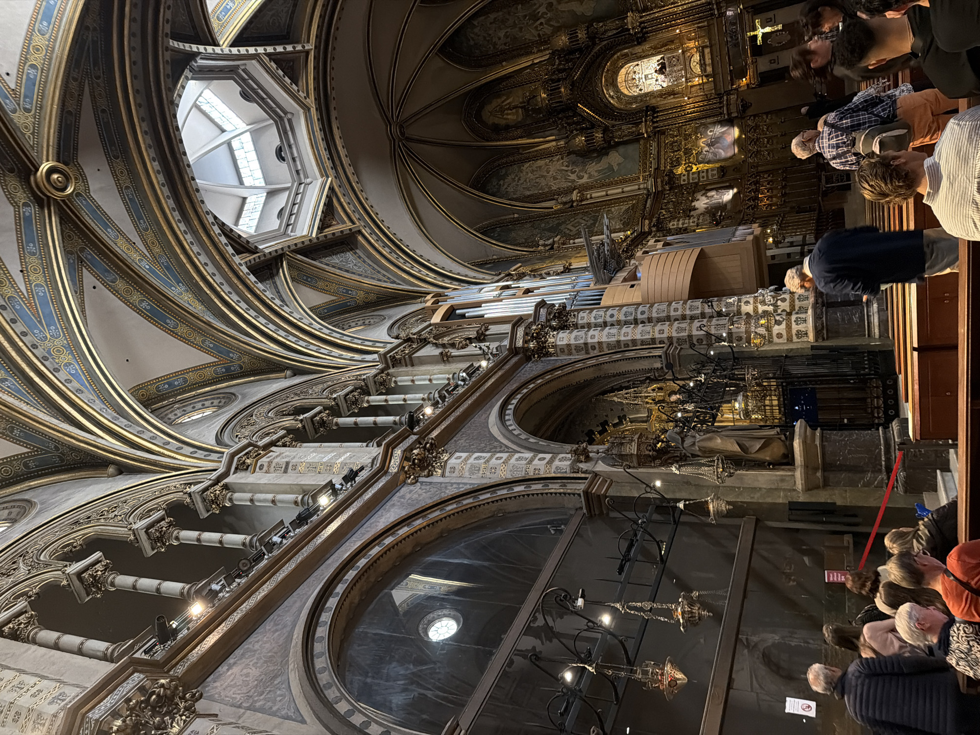 This media file captures the grand and ornate interior of a basilica or monastery, viewed from a low-angle perspective looking upwards. The scene is dominated by the high, vaulted ceilings, which are intricately decorated with elaborate gold and blue patterns on a light background. Majestic ribbed arches curve overhead, leading towards a central octagonal skylight or cupola that allows natural light to enter.
On the right side of the image, a large, complex pipe organ with numerous visible pipes is integrated into the upper architectural levels. Beyond the organ, the sanctuary or altar area is brightly lit and lavishly gilded, featuring religious artwork, statues, and a prominent cross. Ornate columns, balustrades, and decorative balconies line the upper sections of the interior.
In the foreground and lower right, a group of casually dressed people, likely tourists or visitors, are gathered, looking towards the front of the church or upward at the elaborate architecture. A red barrier rope is visible, delineating a path or restricted area for visitors. Decorative chandeliers and hanging lamps provide additional illumination throughout the interior.
The opulent architecture, with its detailed embellishments and prominent pipe organ, suggests a significant religious site, characteristic of locations like the Montserrat Monastery near Vacarisses, Spain. The scene appears to be taking place during the daytime, with a combination of natural light from the skylight and artificial lighting highlighting the interior's rich details. Small, unreadable red informational signs are visible among the crowd.