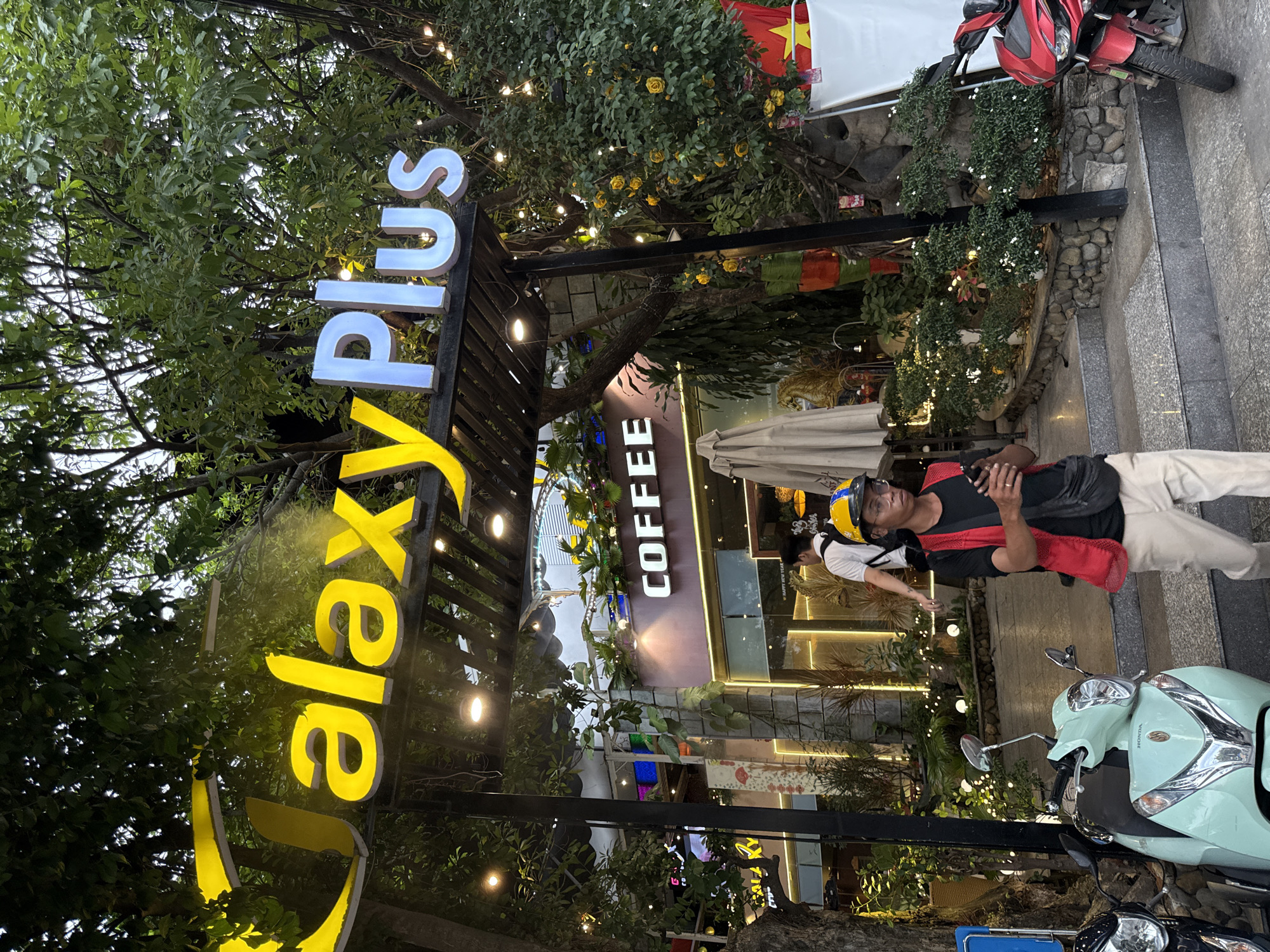 The image depicts a street-level view of "Galaxy Plus," a business located in Nha Trang, Vietnam, indicated by a prominent sign. The sign, featuring yellow lettering for "Galaxy" and white for "Plus," is illuminated and positioned above an entranceway. Below this, a smaller sign reads "COFFEE."
The scene is set outdoors, with lush green trees and foliage surrounding the building, suggesting a tropical environment. String lights are visible, adding to the ambiance. In the foreground, a person wearing a red vest over a black shirt, beige pants, glasses, and a yellow and blue hard hat is walking while looking at their phone. They are carrying a black crossbody bag.
To the left, a light blue Honda scooter is parked, and to the right, a red scooter is partially visible. The ground is paved with stone, and there are steps leading up to the entrance. The background shows the interior of the establishment, with bright lighting and decorative elements, including plants and what appear to be display screens. A Vietnamese flag is visible on the right side of the image. The overall impression is of a bustling commercial area in Nha Trang.
