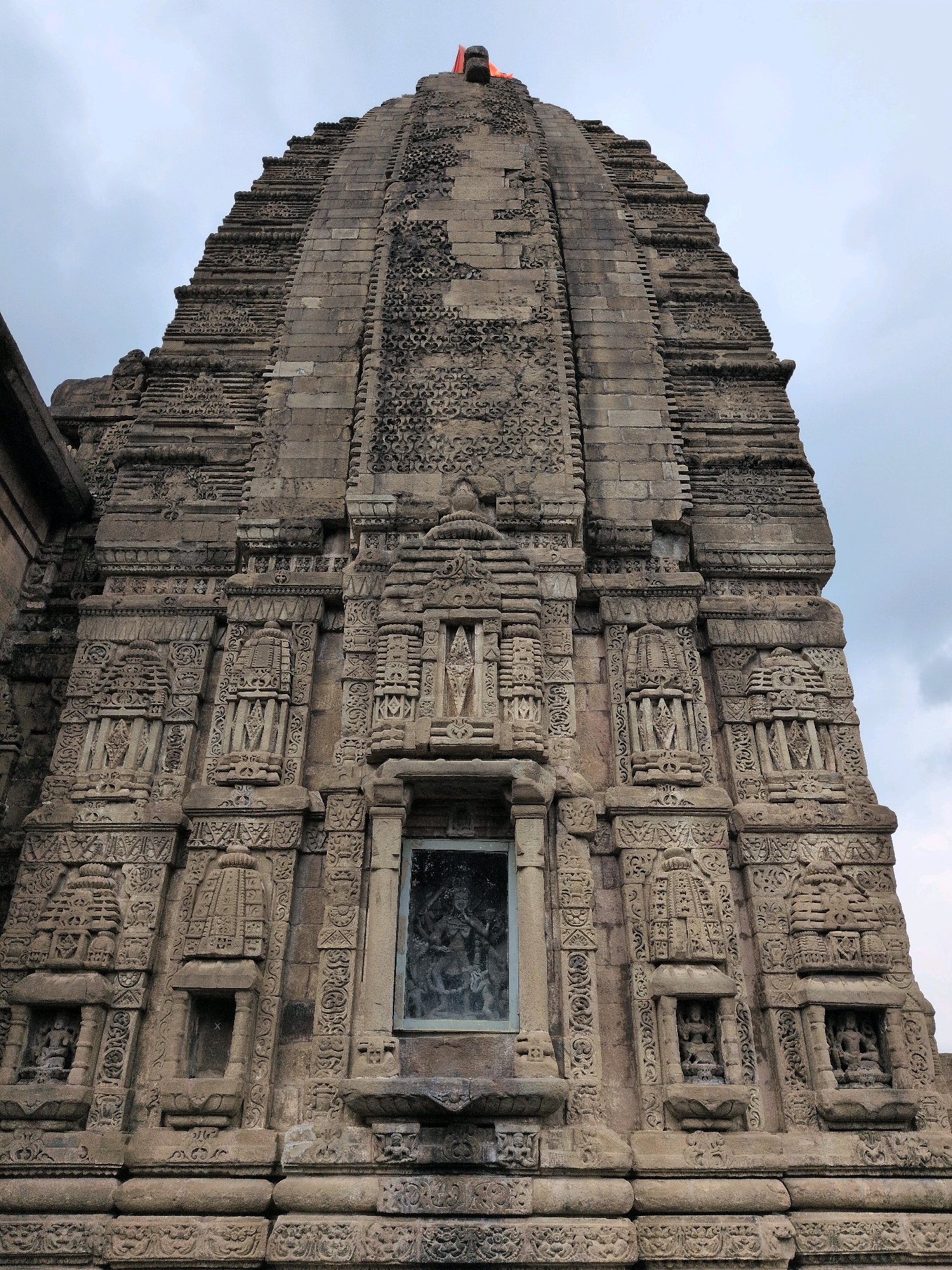 The image shows a low-angle, close-up view of the intricately carved stone superstructure (shikhara) of the Baijnath Temple, located in Baijnath, Himachal Pradesh, India. The temple is made of stone and features elaborate decorative carvings, including miniature temple replicas and sculpted figures. In the center of the facade, there is a large, rectangular window or niche containing a dark, shadowy sculpture of a deity. The sky above is overcast with scattered clouds.