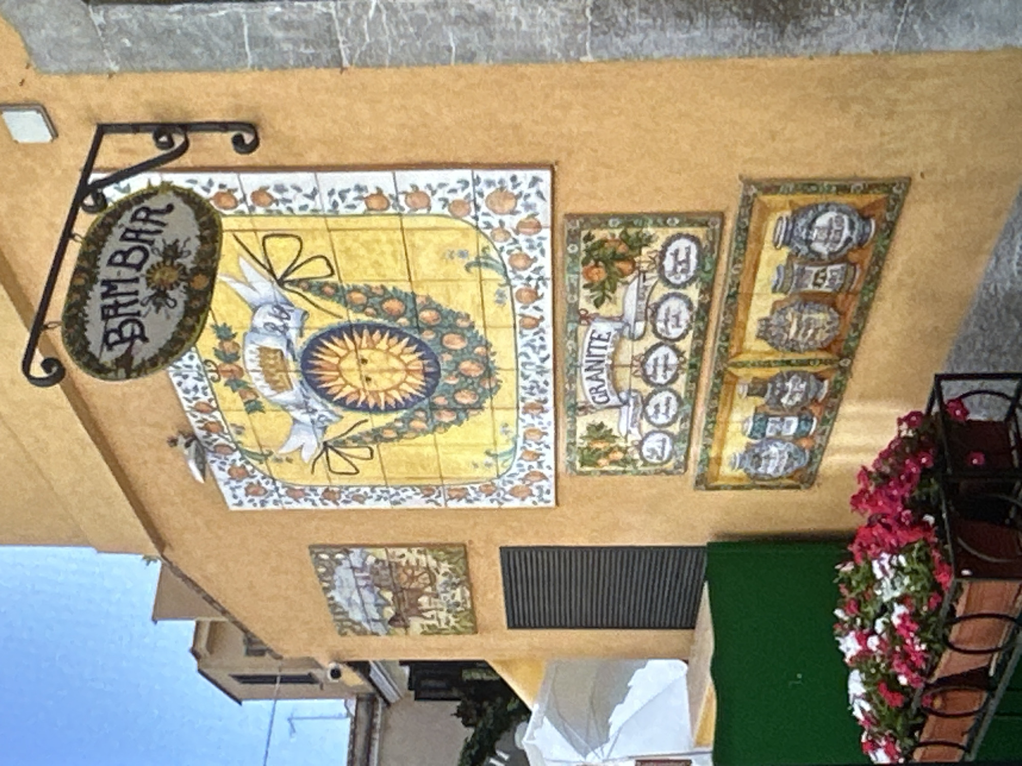 The image shows an outdoor scene with a close-up view of a building facade adorned with decorative tiles. The primary subject is a decorative tile mural on a yellow wall. The mural is colorful, featuring a sun in the center with rays, surrounded by fruit like oranges, and two birds. The sign hanging from an ornate metal bracket above the mural reads "BAM BAR" in an oval shape. To the right of the main mural are two smaller, vertical tile panels. One panel depicts oranges and the word "GRANITE" with circular emblems. The other vertical panel shows a series of urn-like objects. Below the main mural, a smaller square tile panel shows a scene with buildings and trees. A dark, slatted ventilation grate is visible on the wall. To the far right, a planter overflowing with red and white flowers adds a vibrant touch to the scene. The overall impression is one of a charming, possibly Mediterranean, establishment. The lighting suggests it is daytime, with no strong shadows indicating a specific time. The weather appears clear and bright, reflected in the blue sky visible at the bottom left of the frame.