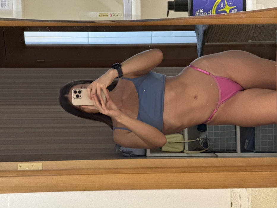 A slender woman in a blue top and pink thong poses for a mirror selfie, holding an iPhone. She is wearing a smartwatch on her left wrist. Behind her, shelves hold various items, including a small, tan purse. The mirror’s wooden frame is visible, reflecting the room’s neutral tones.