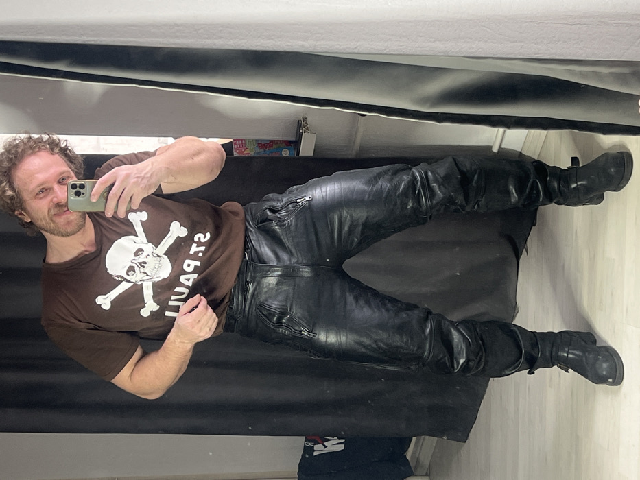 An adult male with light brown, curly hair and a beard is captured in a full-body mirror selfie. He smiles directly towards the camera, which he holds in his right hand. He is wearing a brown, short-sleeved T-shirt featuring a white skull and crossbones emblem with the mirrored text "ST. PAUL" beneath it. He is also dressed in black leather pants with visible zippers on the upper thighs and black leather boots with buckle closures. His left hand rests near his waist. The immediate background on the left is a dark fabric or curtain, partially obscuring a dark item with white lettering. To the right, beyond the mirror's frame, a light-colored shelf holds several small, colorful objects. The floor is light-toned. The specific location within the city of Saint Petersburg, Russia, is not identifiable from the image content.