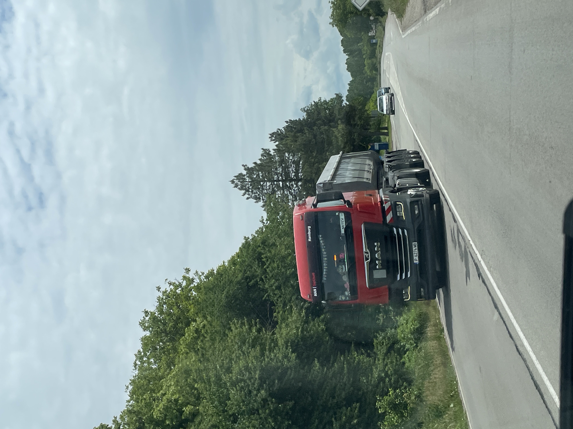 A photograph taken on a roadside in Tsar Kaloyan, Bulgaria, shows a red MAN truck driving on a gray asphalt road, with lush green trees lining the road's edge on both sides. A white vehicle is visible further down the road. The sky overhead is cloudy. The truck's license plate is visible as P 6276 MB.