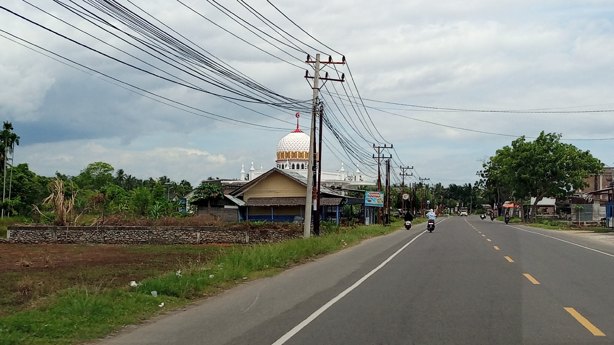 The image shows a road scene in the city of Blangmetimu, Indonesia. A paved road with yellow dashed lines runs through the center of the frame, curving slightly to the right. Several motorcycles are visible on the road, with riders present on some. On the left side of the road, there is an area with dry grass and a low stone wall. Beyond the wall, there is dense green vegetation, including palm trees and other trees. A building with a distinctive white dome, adorned with golden accents and a red crescent moon at the apex, is prominent in the background, suggesting it is a mosque. In front of the mosque, a smaller, older building with a yellow exterior is visible. Utility poles with numerous overhead wires are positioned along the roadside, creating a dense network of lines across the sky. The sky is overcast with grey clouds. On the right side of the road, more buildings and trees are visible, and the road continues into the distance. The overall environment appears to be rural or semi-rural.