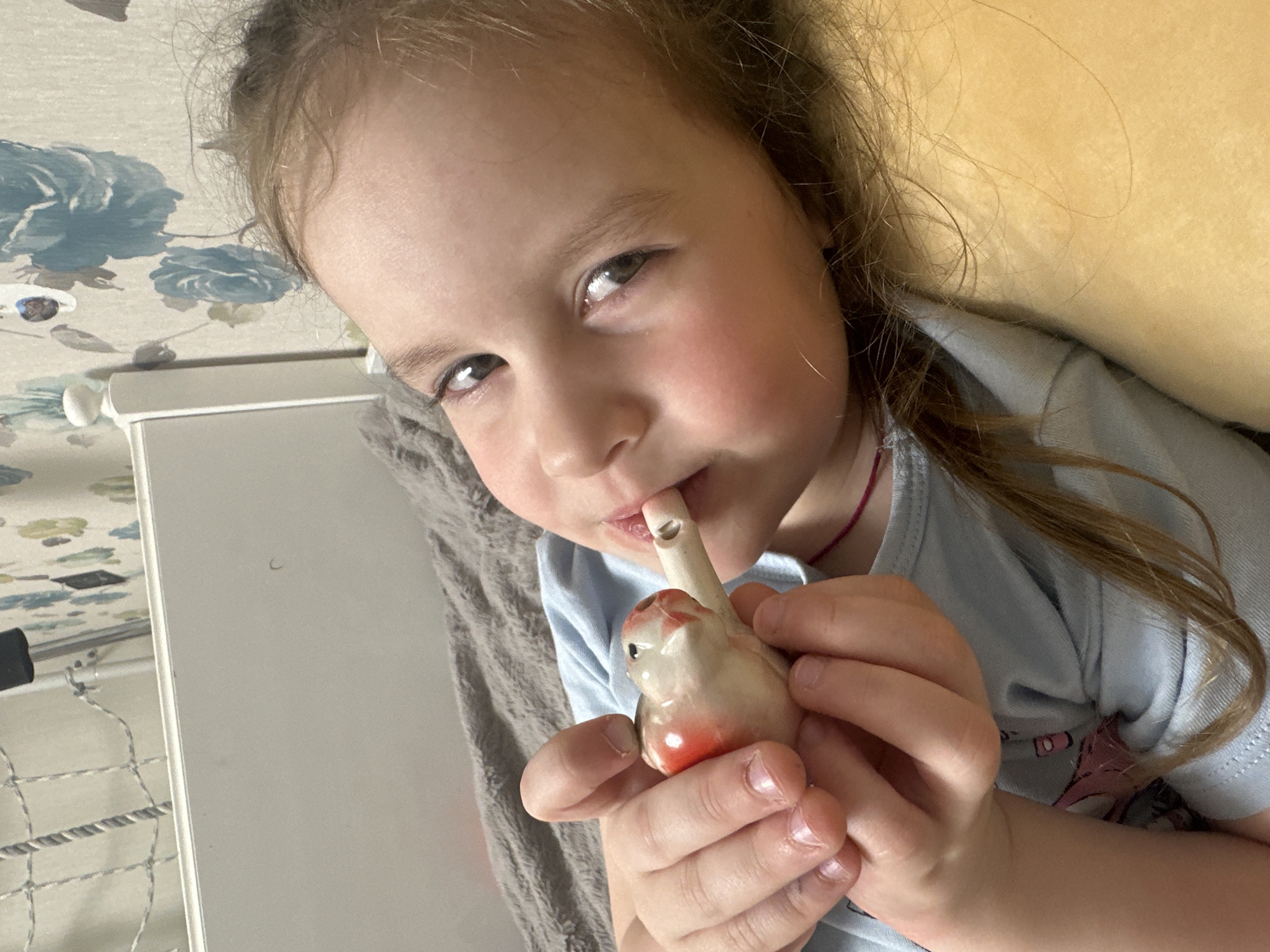 A young girl is holding and playing a bird-shaped ceramic water whistle. She is fair-skinned with brown hair and is wearing a light blue t-shirt. The background includes a floral patterned wall, a white surface, and a grey blanket. The location, Tomilino, Russia, cannot be confirmed from the image alone.