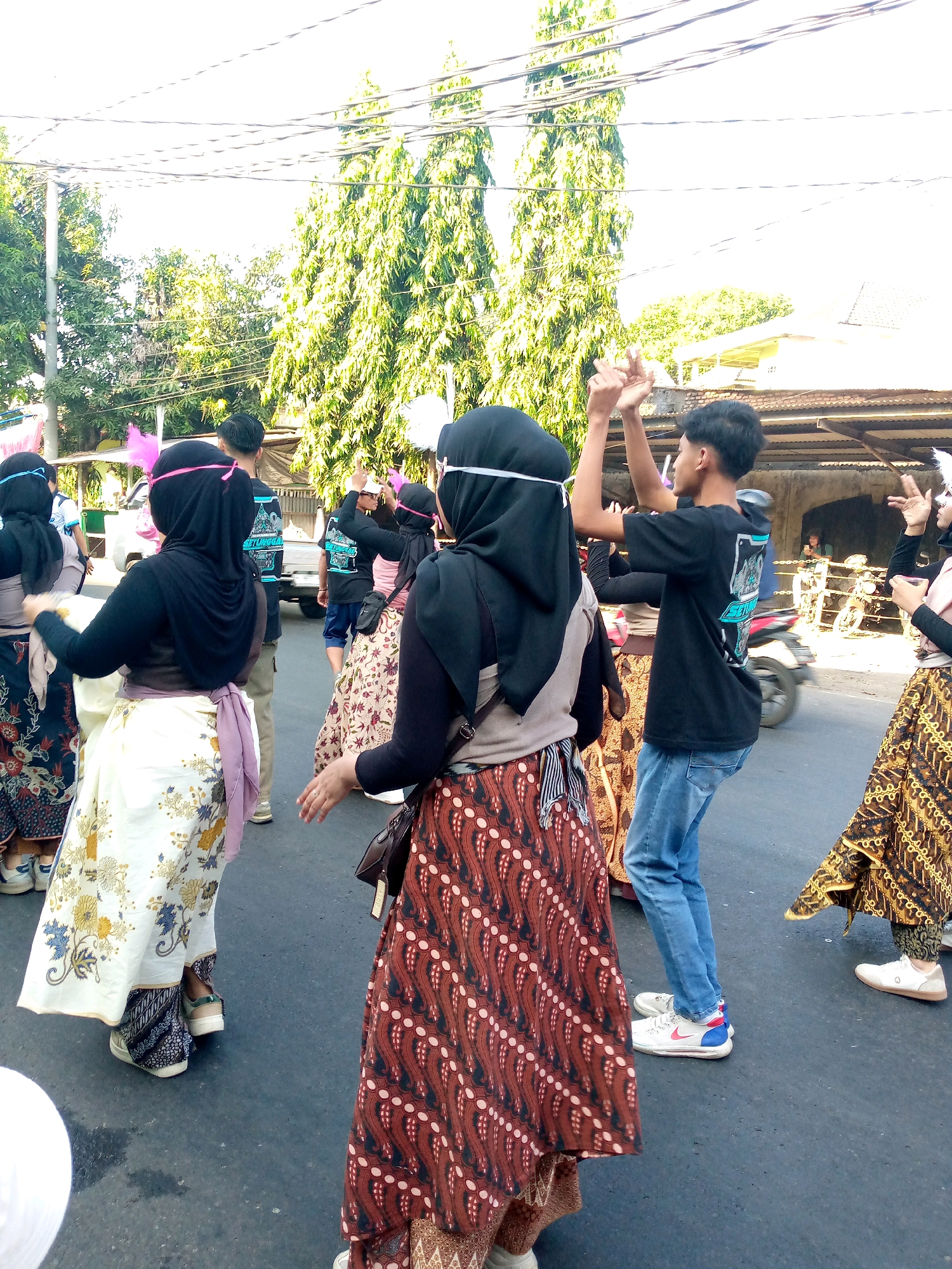 In Sumberwono, Indonesia, a group of individuals, predominantly young women wearing hijabs and batik skirts, are participating in a street performance or parade. The women are adorned with traditional clothing, featuring intricate patterns on their skirts and headscarves, with some accessorizing with pink feathers. They are captured in motion, suggesting a dance or choreographed movement. A young man in a black t-shirt and blue jeans is also present, with his hands raised as if interacting with the group or performing. The background depicts a street scene with trees lining the area, power lines overhead, and a glimpse of buildings and a parked truck. The overall atmosphere appears to be festive and communal, possibly related to a cultural event or celebration.