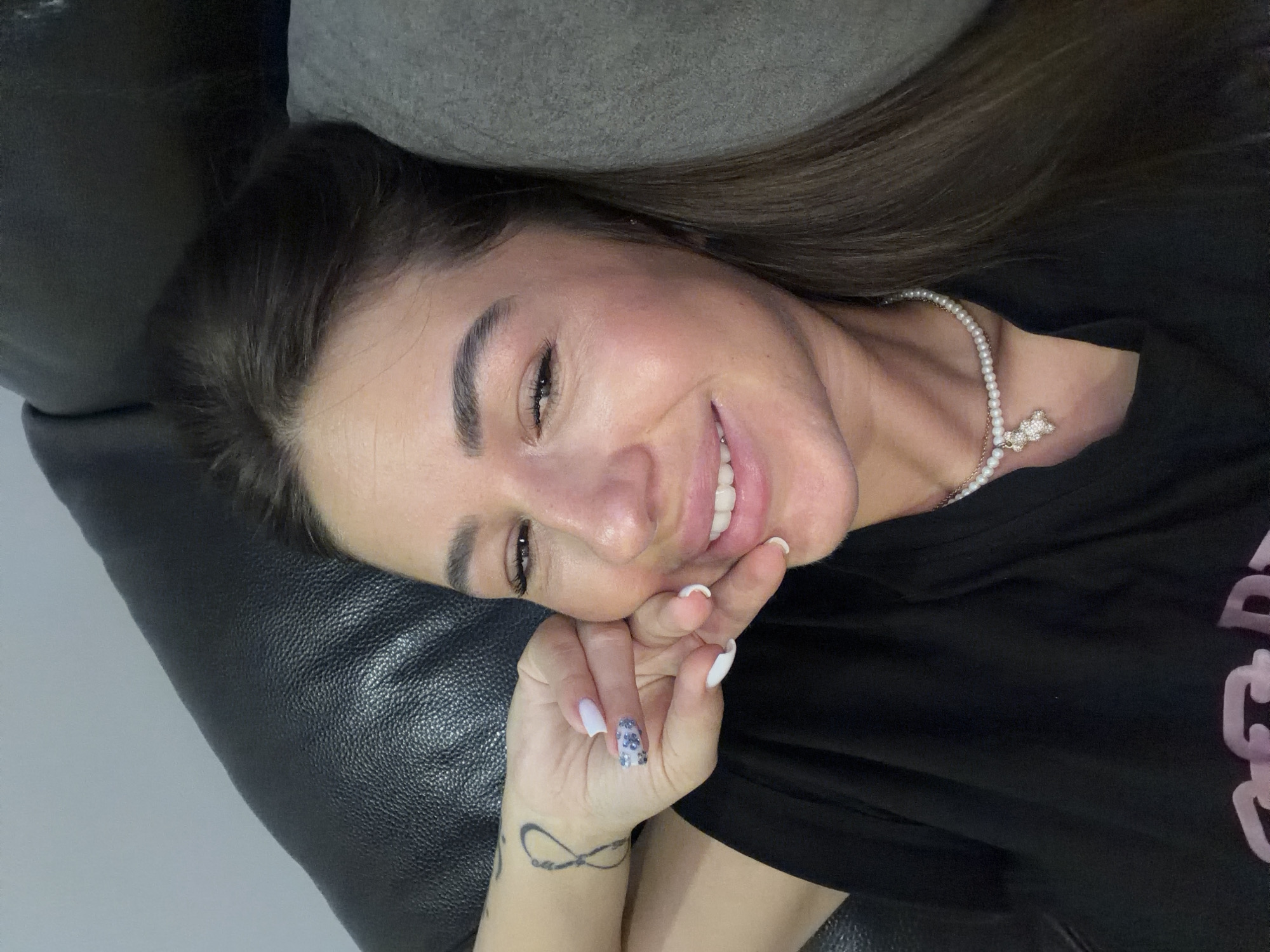 A close-up, eye-level, selfie-style shot captures a woman lying down, resting her head on a dark leather surface and a grey cushion. She is looking towards the camera with a slight smile, her right hand touching her cheek. Her left arm is visible, featuring an infinity tattoo on the wrist and manicured fingernails with a white and sparkly design. She is wearing a black t-shirt and a pearl necklace with a diamond-encrusted teddy bear pendant. The background is indistinct, suggesting an indoor setting. The image does not contain any information about the city of Novi Iskar, Bulgaria.