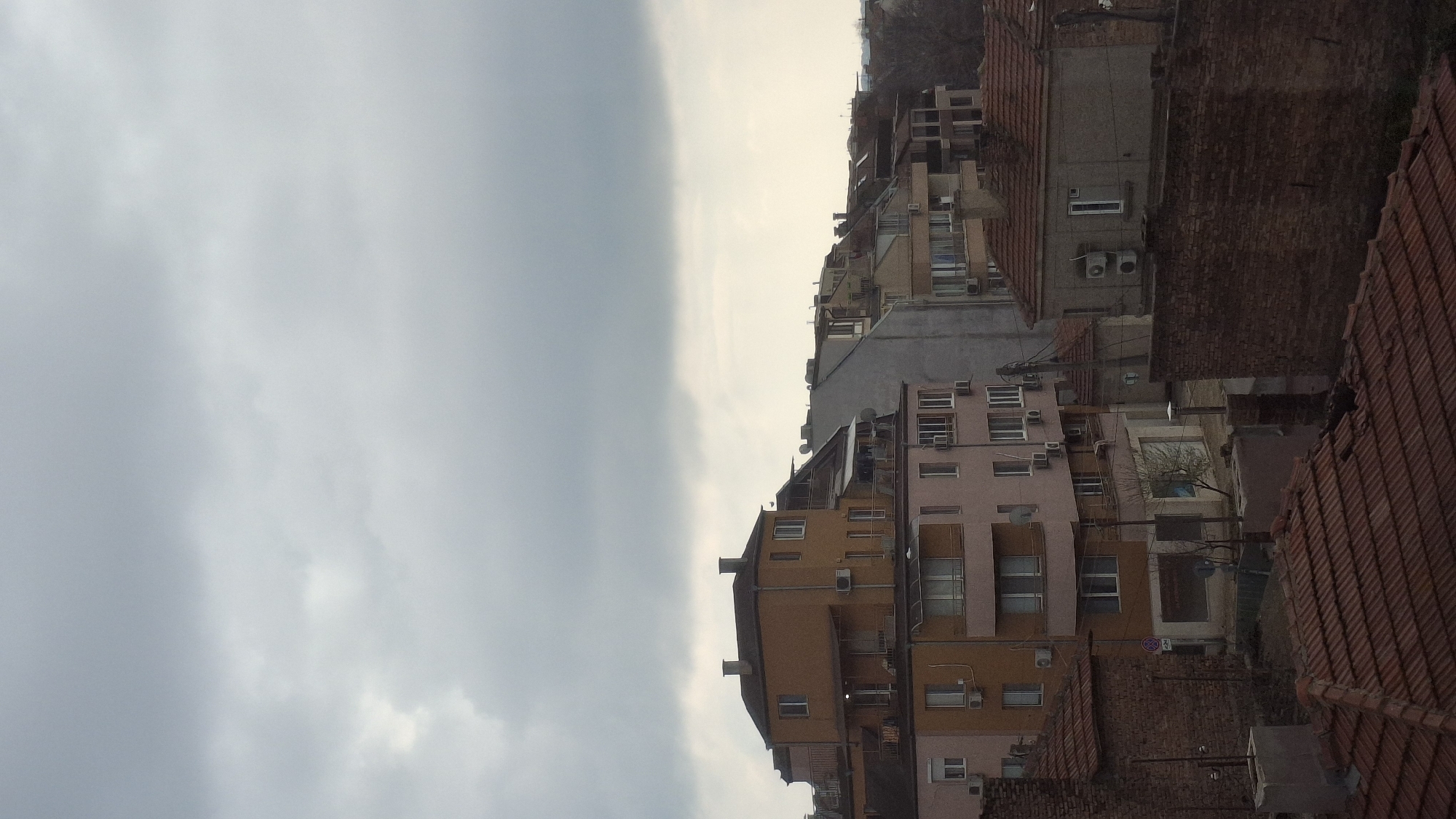 The image depicts a densely built urban landscape, likely situated in Varna, Bulgaria. The architecture is characterized by a mix of residential buildings, predominantly medium-rise, with varied colors ranging from pink and brown to grey. Many of the buildings feature external air conditioning units. Red tiled roofs are prominent. The sky is overcast, with a mix of grey and white clouds. The perspective is somewhat elevated, suggesting the photo was taken from a higher vantage point. There are no visible people.