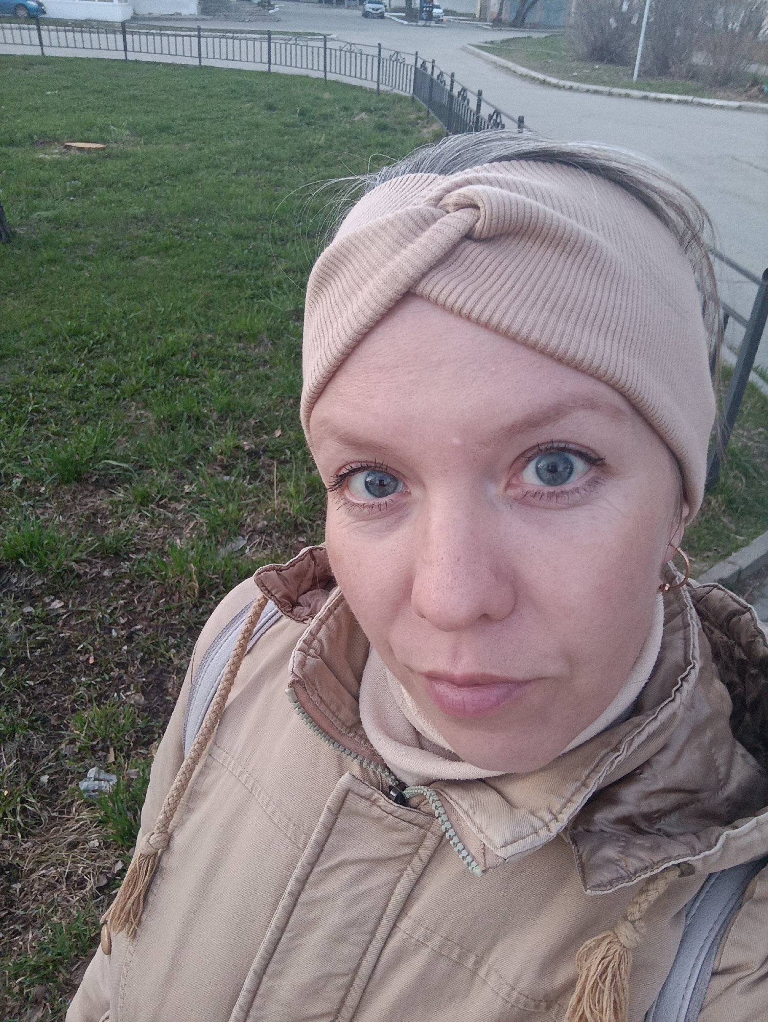 A head-and-shoulders selfie captures a woman outdoors in Chaikovsky, Russia. She looks directly into the camera with wide blue eyes and a neutral, slightly inquisitive expression, her mouth slightly ajar. She wears a light beige ribbed headband with a twisted knot design on her forehead, and her gray hair is visible around it. Her attire includes a warm-looking tan or beige winter jacket with a prominent zipper and braided rope details, worn over a light-colored turtleneck or high-collared garment. A light-colored backpack strap is visible over her right shoulder, and a small gold-colored hoop earring adorns her right ear.
The background shows an urban or suburban environment. Immediately behind the woman is a grassy area with some patches of dry earth and a small tree stump. A dark metal fence separates this green space from a curving asphalt road or sidewalk. In the distance, bare trees or bushes line the road, and indistinct buildings are visible. A blue car is parked on the road in the far left background, and another vehicle is partially seen on the right. The soft, diffused lighting suggests it is late afternoon or early evening. The woman's warm clothing and the bare vegetation indicate cool weather, consistent with late autumn, winter, or early spring. There is no visible text in the image.