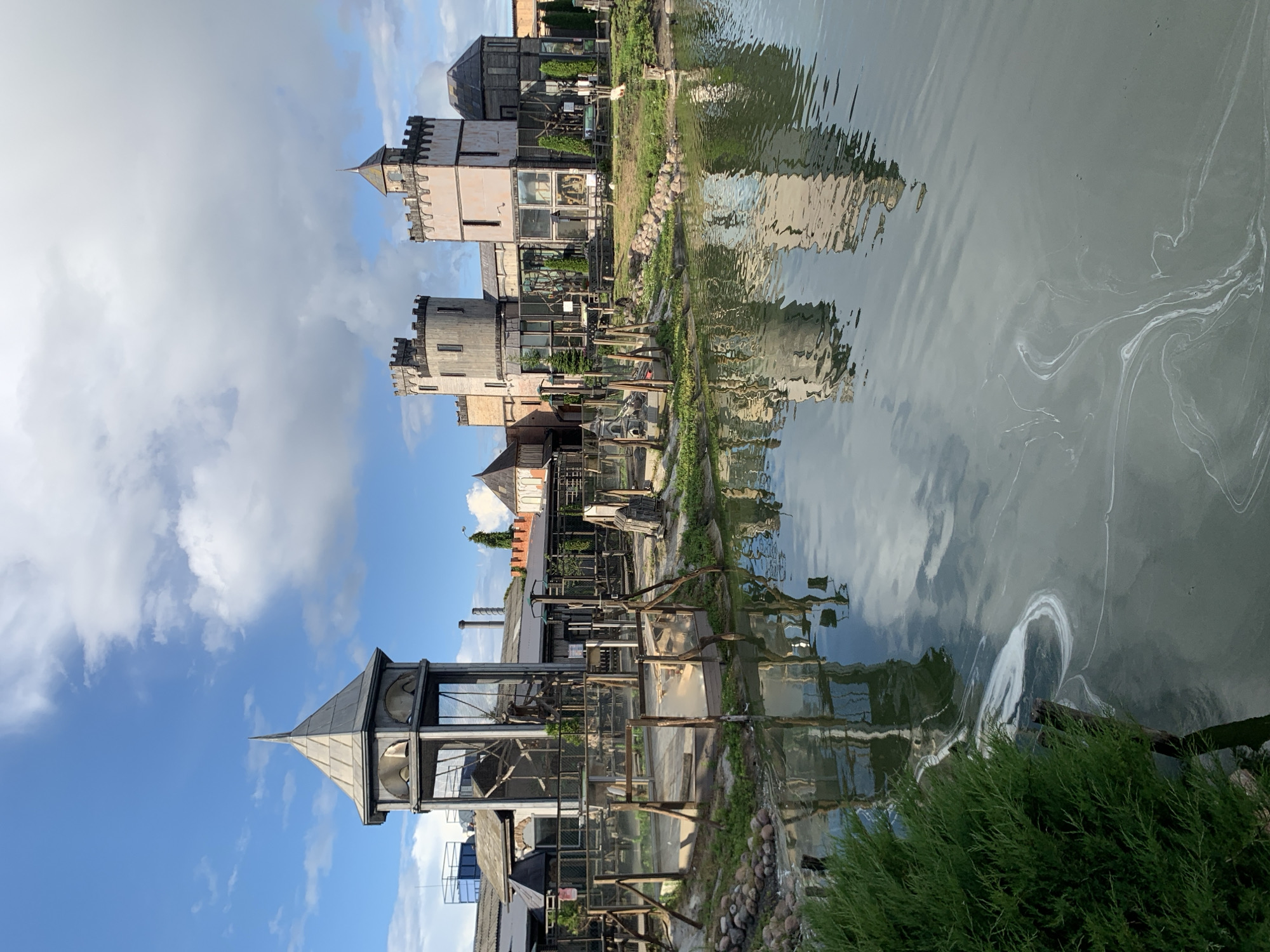 The image shows a daylight, exterior view of an amusement park or attraction featuring a castle-like structure and a water feature. The main structure is a series of interconnected buildings with towers and crenellations, resembling a medieval fortress. One prominent tower has a pointed, conical roof. The buildings appear to be constructed with a mix of materials, including stone, brick, and glass. In front of the castle structure is a body of water, possibly a pond or small lake, with reflections of the buildings visible on its surface. There are also some wooden fences and enclosures in the foreground, suggesting animal exhibits or play areas. Greenery, including trees and bushes, is present around the water and the structures. The sky is blue with scattered white clouds. The overall scene suggests a recreational or entertainment venue designed with a historical or fantasy theme. The provided location "Малахавецкі сельскі Савет, Belarus" indicates this is a rural area in Belarus.