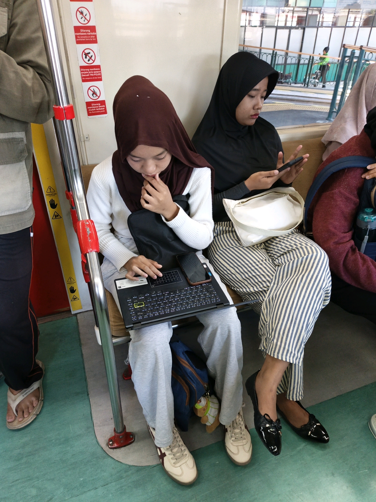 The image depicts an interior scene, likely on a ferry or train, with several individuals present. In the foreground, a person wearing a brown hijab and a white sweater is seated, looking down at a laptop with a keyboard. To their right, another individual in a black hijab and striped pants is also seated, holding a smartphone. A white bag rests on their lap. A third person, partially visible on the left, wears dark pants and flip-flops. The environment includes a metal pole with red fixtures and warning signs attached to a wall. The flooring is a muted green color. In the background, a window offers a view of a station platform with other individuals and structures. The atmosphere suggests a public transportation setting, possibly in the city of Klaten, Indonesia, given the warning signs in Indonesian that include prohibitions against weapons and narcotics.