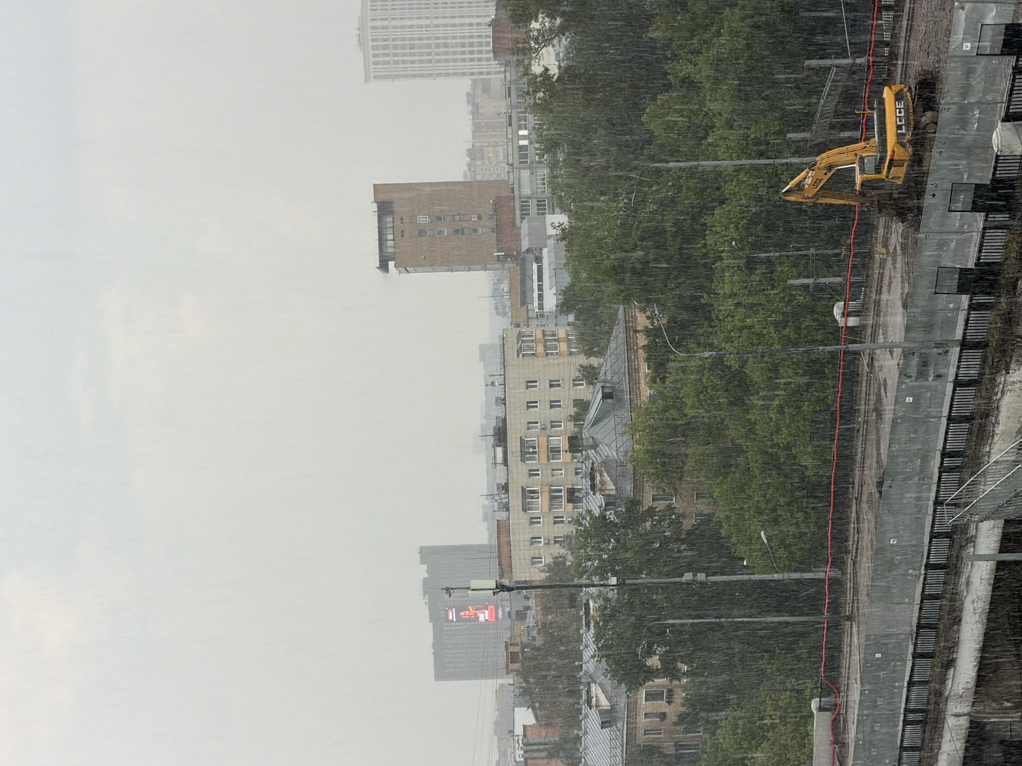 This is a medium shot, taken from a high vantage point, of a city landscape during a heavy rainfall. The sky is overcast and a pale grey. In the middle ground, several multi-story buildings are visible, with varying architectural styles, some appearing residential and others more office-like. Trees with dense green foliage are interspersed between and behind the buildings. In the foreground, a construction site is visible with a yellow excavator on a muddy surface. A red rope runs across the construction area, and a black metal fence separates it from a walkway or road. Streetlights are visible throughout the scene. The rain is heavy, with visible streaks falling throughout the image, creating a somewhat hazy effect. The overall impression is one of urban construction and city life being impacted by inclement weather. While the specific location is not definitively identifiable from the visual alone, the architectural styles and the presence of Cyrillic text on one of the buildings (partially obscured but resembling Russian lettering) suggest a location in Moscow, Russia.