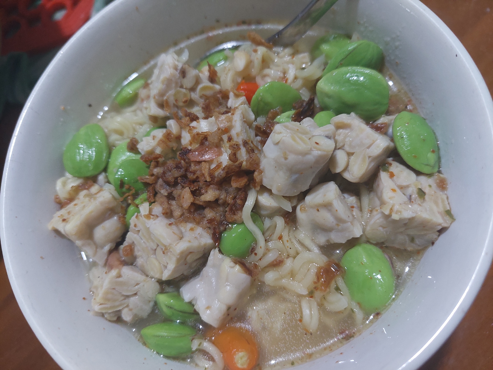 This is a close-up, overhead shot of a bowl of food. The dish appears to be a type of soup or stew with noodles, cubed white pieces that could be tofu or tempeh, bright green pod-like vegetables, and topped with fried shallots. There are also small pieces of red chili visible. The bowl is white and sits on a wooden surface. The background is blurred but suggests an indoor setting. The specific location in the city of Jelutung, Indonesia, cannot be determined from the image alone.