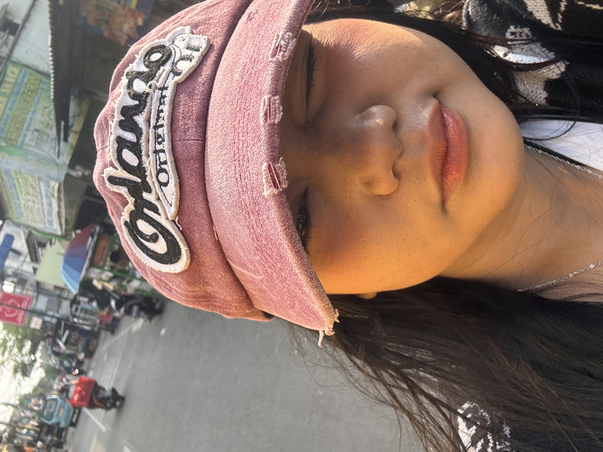 The image shows a close-up of a young woman wearing a pink baseball cap with "Orlando Original" embroidered on it. She is looking to the side, with one eye squinting. The background is blurred, but appears to be a street scene in North Jakarta, Indonesia. There are buildings, shops, and vehicles visible, including a blue van and several motorbikes. The lighting suggests it is daytime, possibly late afternoon, with the sun casting a warm glow on the woman's face. Her dark hair is visible, and she is wearing a necklace. The overall impression is of a casual, outdoor setting within an urban environment.
