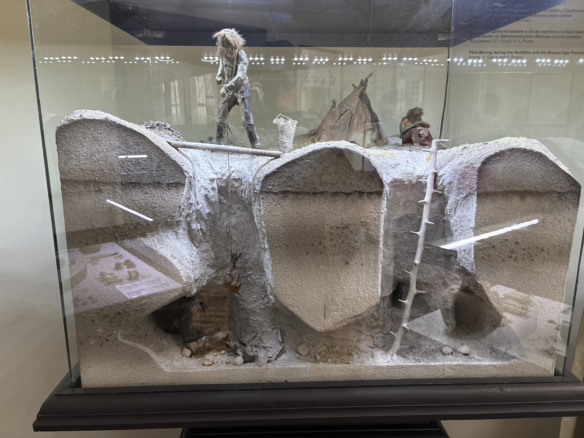 The image displays a diorama within a glass case, depicting a scene of flint mining during the Neolithic and Bronze Age periods. The diorama is sectioned to reveal subterranean activity, showing layered soil and rock formations. In the upper portion of the scene, a figure, appearing to be a representation of an early human, stands near a crude tent structure. This figure is shown actively engaged in mining, with one hand holding what appears to be a tool, and the other holding a rope that leads down into one of the excavated areas. Beside the figure is a woven basket, likely for carrying materials. Another seated figure, also resembling an early human, is positioned further back, near the tent.
The excavated areas reveal tunnels and chambers dug into the earth. One tunnel features a rudimentary ladder made of branches, providing access to a lower level. Within these excavated spaces, there are representations of unearthed materials and possibly tools or artifacts. The overall environment of the diorama is earthy and rough, with textures simulating stone, soil, and vegetation.
A text label is visible at the top of the display case. Part of the English text reads: "Flint Mining during the Neolithic and the Bronze Age Periods. Artists: V. Golub and F. Rozov." The diorama is situated in the city of Hrodna, Belarus.