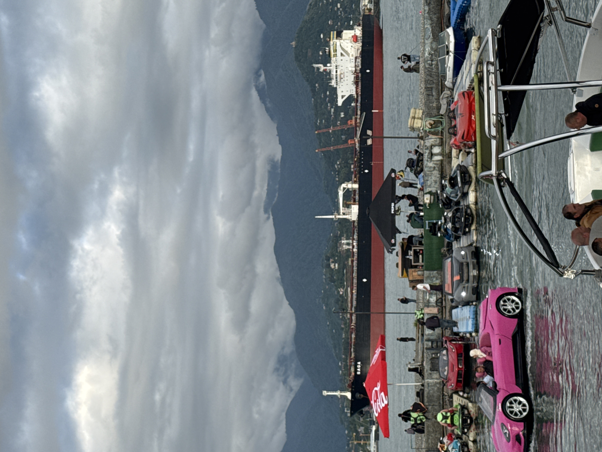 The scene is set in Batumi, Georgia, on a pier with a large cargo ship docked nearby. The sky is overcast with heavy clouds. The pier is bustling with activity, featuring a display of miniature vehicles, including a bright pink one prominently in the foreground. People are gathered around, observing the display and the surroundings. A large red Coca-Cola banner flutters in the breeze. Boats are docked along the pier, with people visible on board. In the background, a mountain range looms under the cloudy sky.