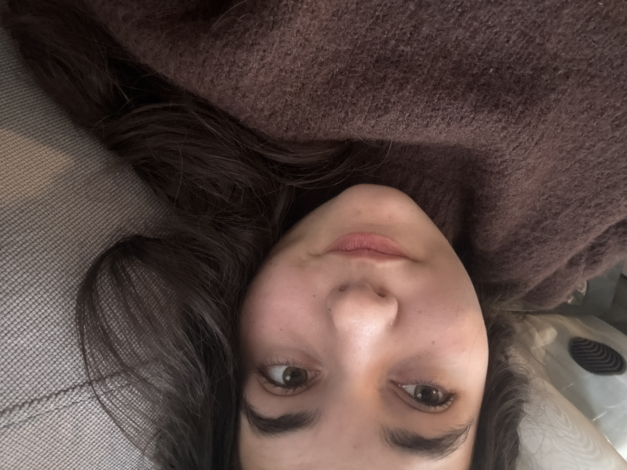 The image is a close-up shot of a woman's face, seemingly taken from an upside-down perspective. She has fair skin, dark brown hair, and brown eyes. The woman appears to be wearing a brown, textured sweater or garment that fills a large portion of the upper part of the image. To her left, there is a grayish, textured surface. The background contains a hint of a rounded, white or light-colored object. There is no indication of the location being Plovdiv, Bulgaria, as there are no geographical landmarks or culturally specific objects visible in the frame.