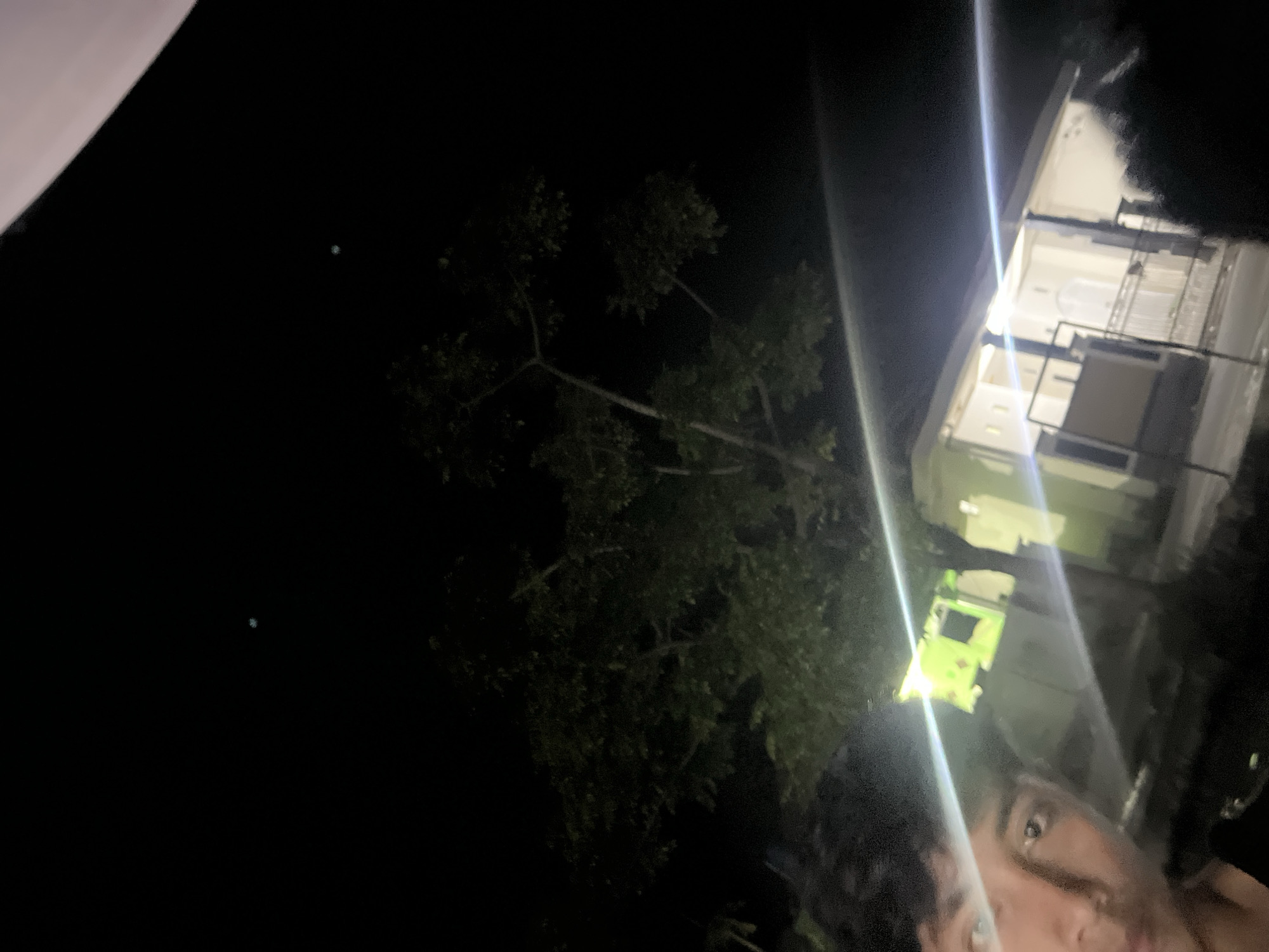 The image is a night-time selfie taken outdoors, likely in Laban, Indonesia. A portion of a person's face is visible in the foreground on the left, with their eyes looking towards the camera. The background is dark, with a tree and a house illuminated by artificial lights. The house appears to be a single-story structure with windows and a covered porch. There are also bright streaks of light across the image, possibly from light sources or a camera artifact. The sky is black with two faint, blurry lights visible in the upper portion of the image.