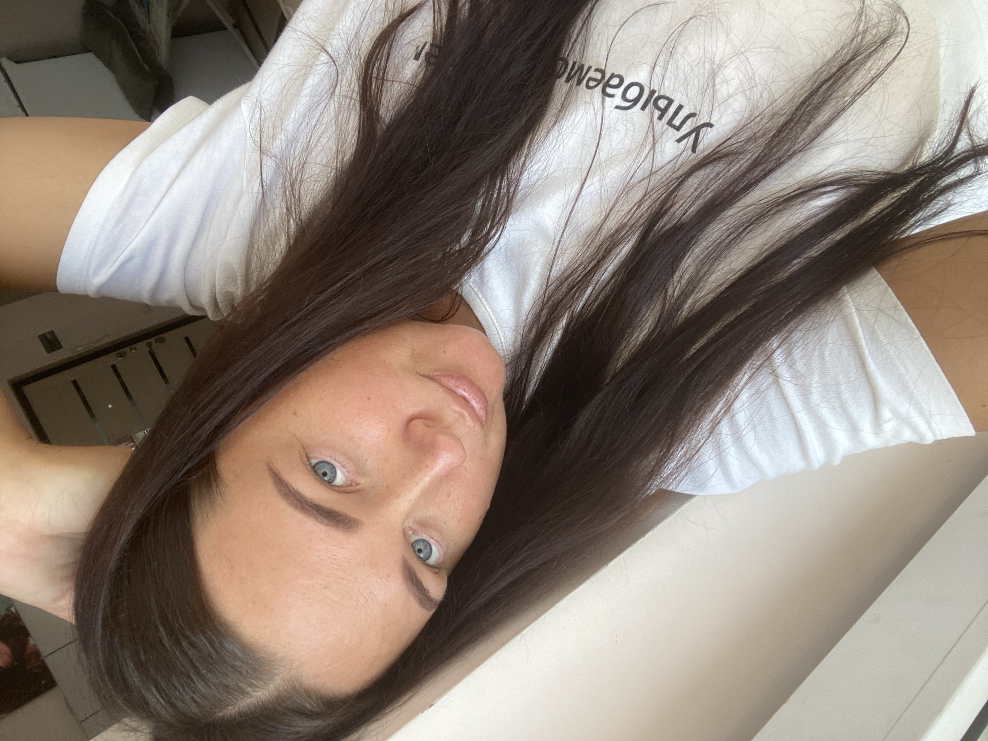 A woman with dark brown hair and blue eyes, wearing a white t-shirt with Russian text, takes a selfie. Her right arm is raised above her head, and her hair is falling around her face and shoulders. The background appears to be indoors, with a light-colored wall and a glimpse of furniture. The text on her shirt reads "Улыбаемся," which translates to "We are smiling."