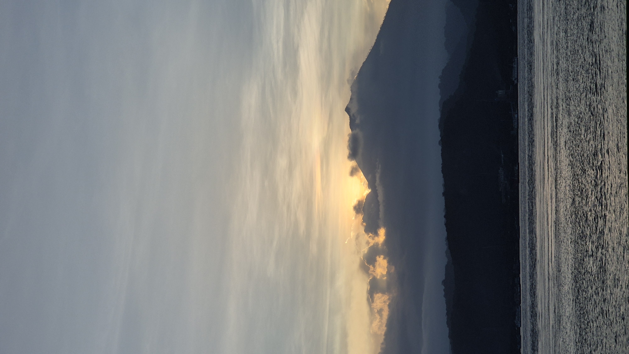The image shows a landscape at sunset or sunrise, likely near water and mountains. The sky is filled with clouds tinged with orange and yellow light from the sun, which appears to be partially obscured by the mountain range in the center of the frame. The mountain range is dark, with some cloud cover around its peaks. To the right of the mountains, there is a body of water that is reflecting the light, creating vertical patterns. The location is presumed to be Unknown, Indonesia. No people are visible.