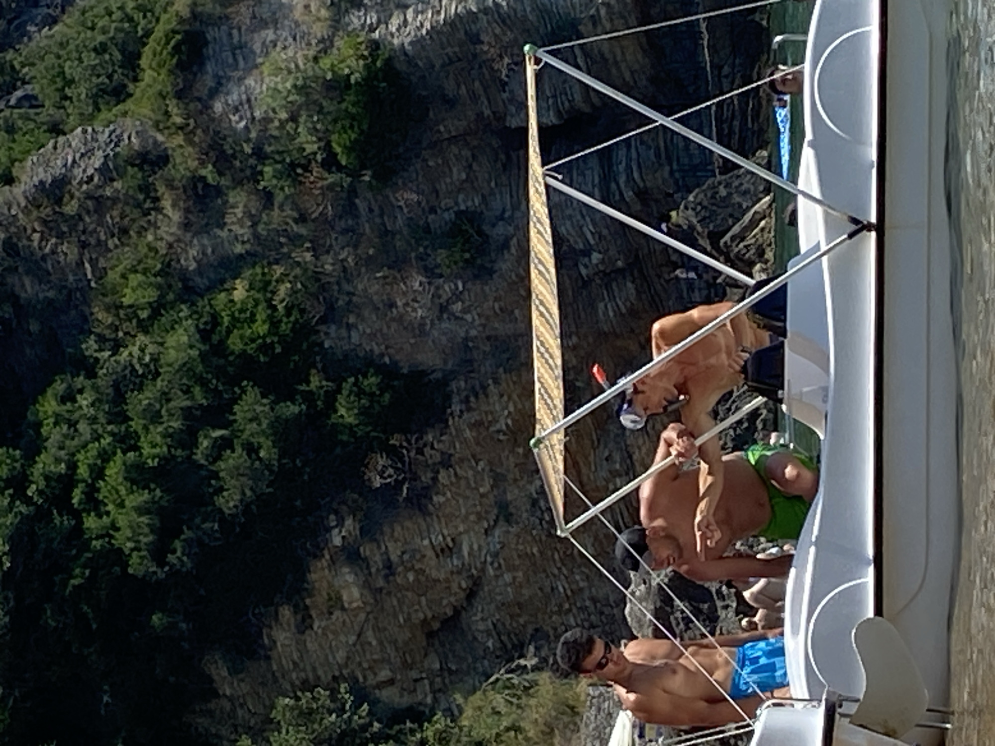 Three men are on a white pedal boat near a rocky, tree-covered cliff face, likely in Budva, Montenegro. The man on the far left, wearing blue swim trunks and sunglasses, stands looking forward. In the middle, a man with a black cap and green swim trunks leans over the side of the boat, gesturing with his right hand towards the water. To his right, a man wearing a snorkel mask and a black top appears to be interacting with him. The water is clear and calm, with sunlight glinting off its surface. The rocky cliff rises steeply behind the boat, covered with dense green foliage. The scene captures a moment of leisure and outdoor activity on a sunny day.