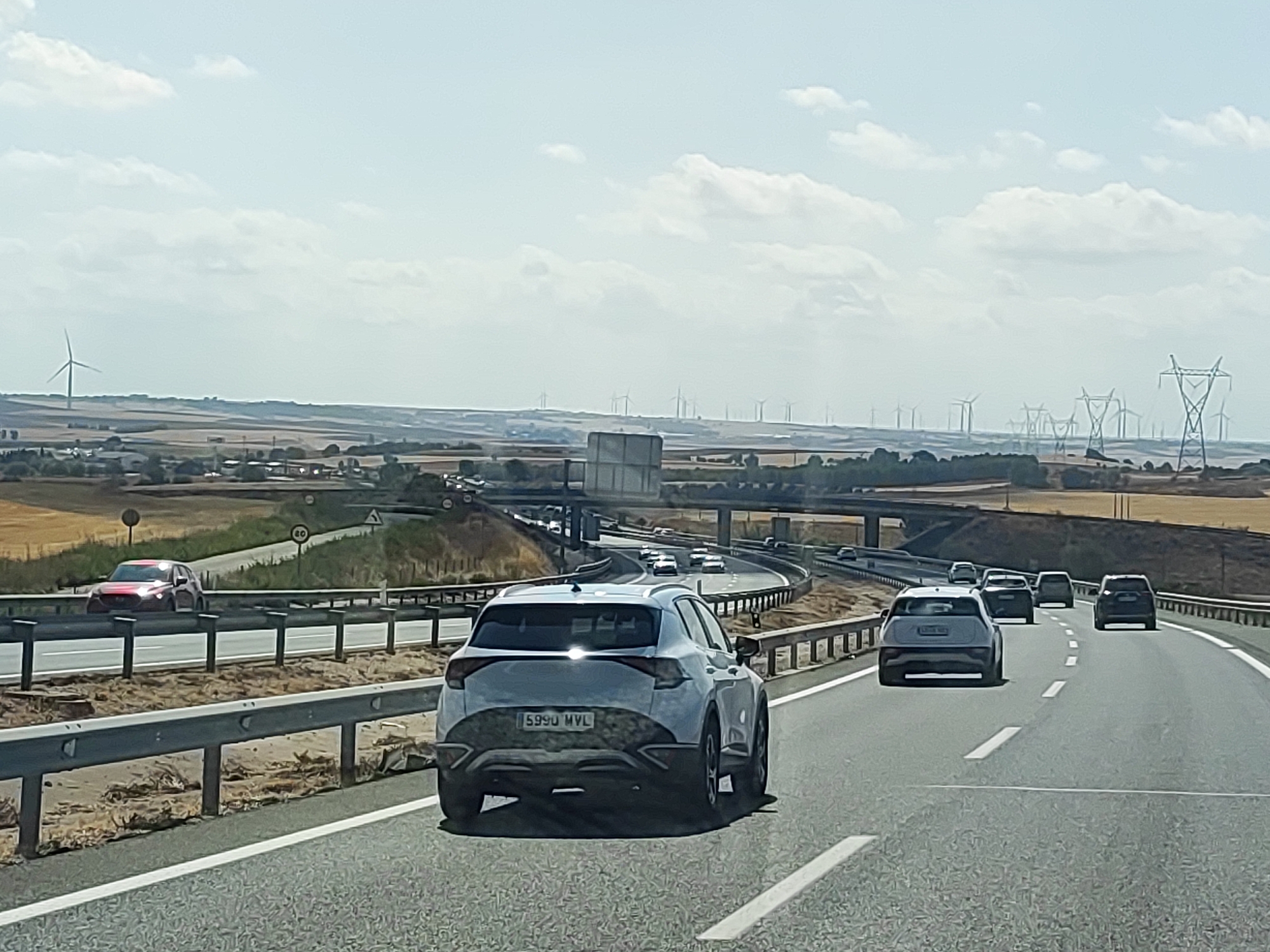The image shows a highway scene on a clear, sunny day, likely in the vicinity of Castillo de Garcimuñoz, Spain, characterized by a flat, rural landscape with scattered wind turbines and power lines in the distance. The foreground features a white SUV with a license plate reading "5990 MVL" driving on the right side of the road. Several other vehicles are visible on the highway, including a red SUV in the left lane and a line of cars further ahead. The highway has multiple lanes separated by white lines and is bordered by a metal guardrail and a dry, sandy embankment. The landscape beyond the highway consists of agricultural fields, some appearing to be harvested, and a few small buildings in the far distance. A pedestrian overpass is visible in the mid-ground. The sky is blue with scattered white clouds.