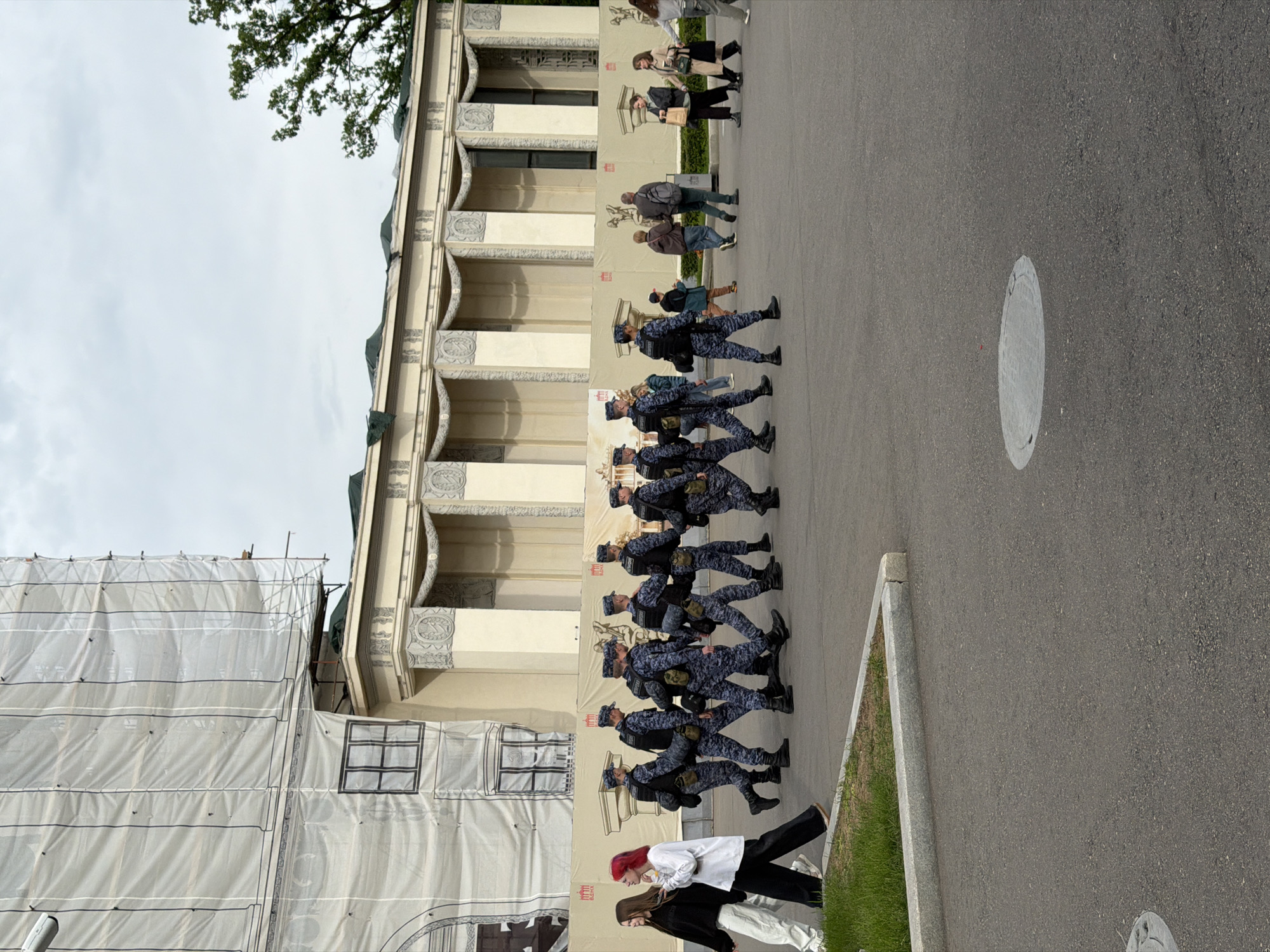 A group of military personnel in blue camouflage uniforms march in a line along a paved road. The individuals are wearing caps and appear to be carrying equipment on their backs. Beside them is a building undergoing renovation, covered in protective sheeting. People are walking near the troops, including one person in a white shirt and black pants. The road is asphalt, and the sky is overcast. The location is Moscow, Russia.