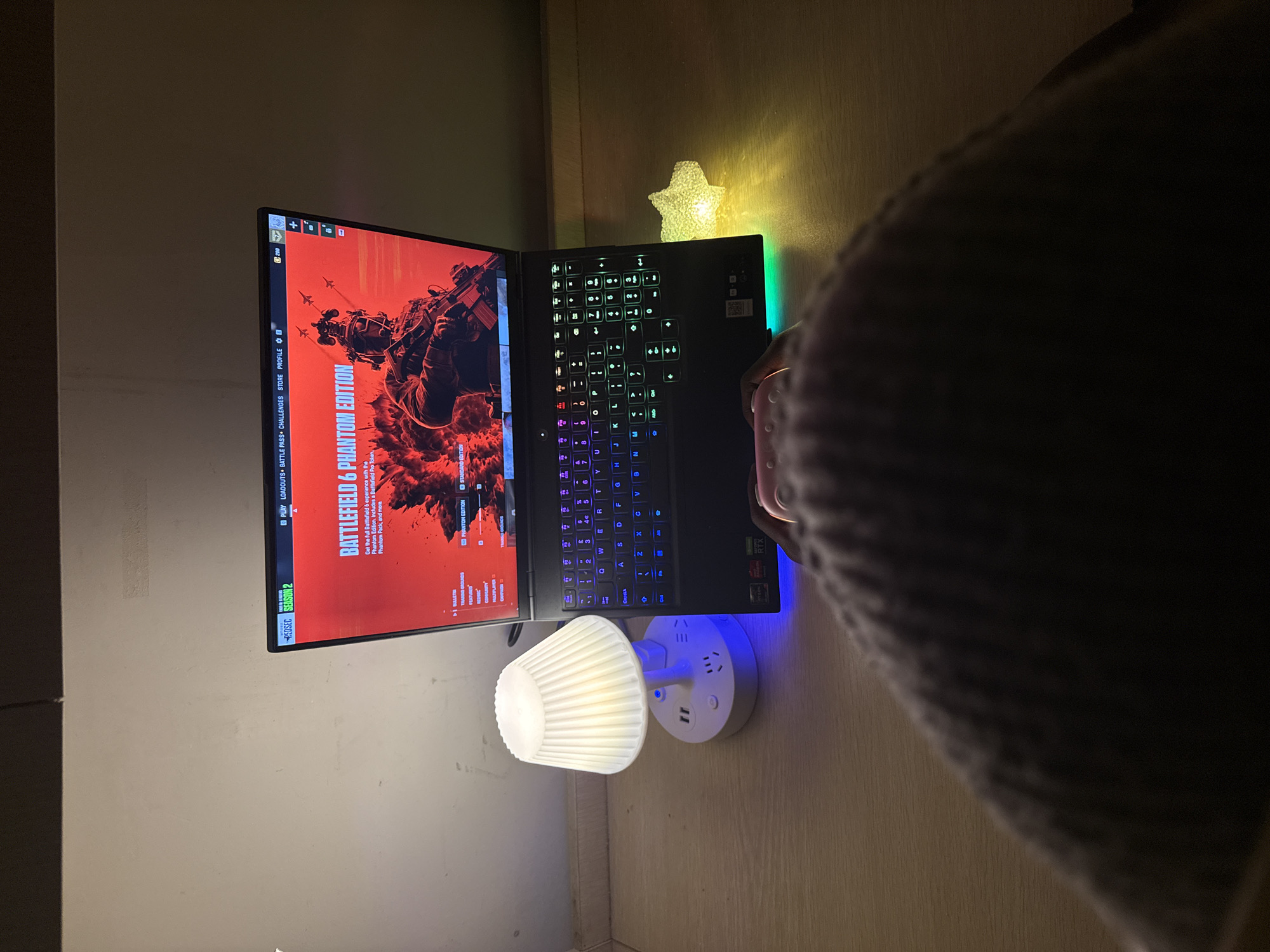 A laptop is open, displaying the game "Battlefield 6 Phantom Edition" on the screen. The keyboard of the laptop has colored backlighting. Next to the laptop, there is a nightlight with a white lampshade attached to a white outlet adapter that has USB charging ports. Above the laptop is a small, star-shaped decorative light. The laptop is on a surface that appears to be a desk or table, with a wall behind it. A portion of a person's body, possibly wearing a gray sweater, is visible on the right side of the image. There is no indication of the location of Jiangjin, China.
