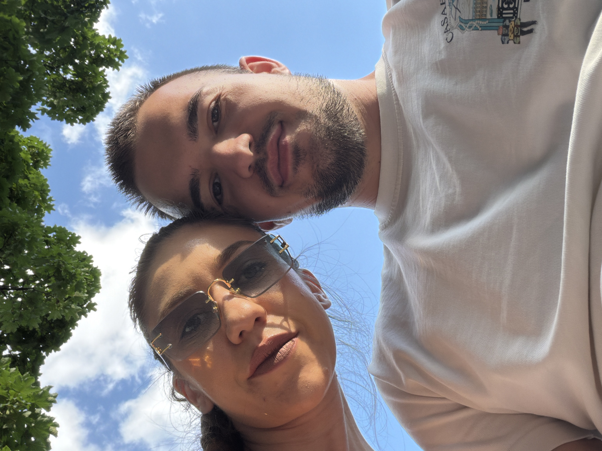An outdoor, low-angle selfie captures two individuals, a female and a male, positioned in close proximity against a bright blue sky with scattered white clouds and green foliage overhead. The scene is illuminated by strong daylight.
The female is on the left side of the frame, looking towards the camera. She has tanned skin, dark hair (partially visible), and is wearing rimless, dark-tinted rectangular sunglasses with gold-colored metal accents on the bridge and temples. A small gold-toned charm dangles from the right lens. Her lips are lightly colored and slightly parted. She is wearing a light-colored (possibly off-white or light beige) top, visible on her upper chest and shoulder.
The male is on the right side of the frame, looking directly at the camera. He has short, dark, spiky hair, a short beard, and tanned skin. He is wearing a light-colored (likely off-white or light beige) crew-neck t-shirt. On the right side of his chest, a graphic illustration is visible, featuring an architectural design with small human figures below it, and the partial word "CASABLANCA" printed above the illustration.
The background consists of a clear blue sky with a few wispy white clouds. Dense green tree leaves are visible at the top of the frame, suggesting the photo was taken while looking upwards, possibly from under a tree.
The location is in the city of Sofia, Bulgaria.