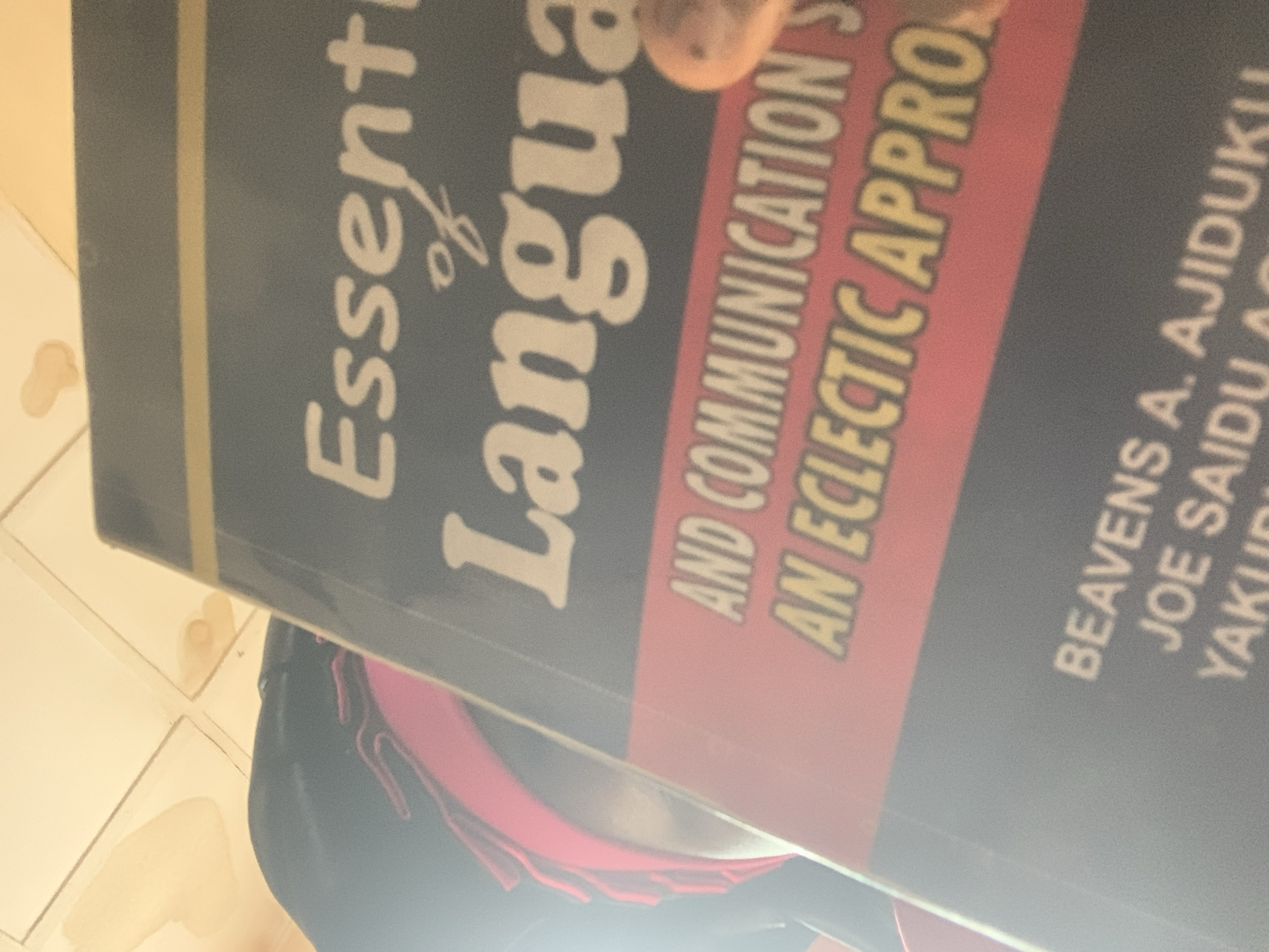 The image shows a close-up of a book titled "Essentials of Language and Communication: An Eclectic Approach" held by an individual. The book's cover is dark gray with white lettering. The author names "BEAVENS A. AJIDUKU" and "JOE SAIDU" are visible at the bottom of the book. The person holding the book is wearing a black headscarf and a pink garment underneath. The background appears to be a tiled surface with some light brown stains. The scene is captured from a low angle, looking upwards. There is no discernible location in Jauro Yinu, Nigeria.