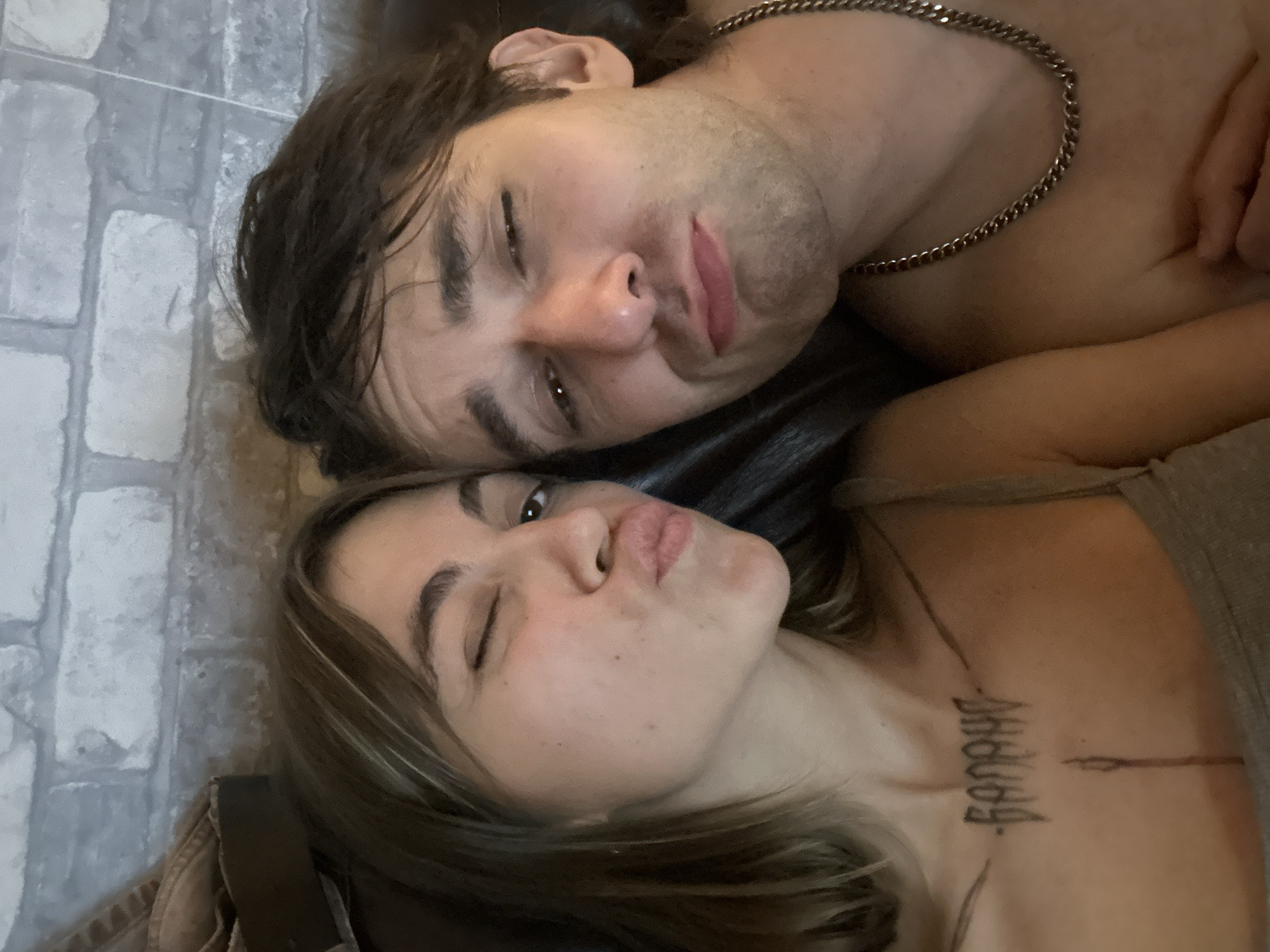 Two individuals, a woman and a man, are positioned in close proximity, facing the camera. The woman, on the left, has brown hair with highlights and is winking, with her lips puckered. She is wearing a grey tank top and has a tattoo on her chest. The man, on the right, has dark, tousled hair and is looking towards the camera with a slightly pouty expression. He is wearing a silver chain necklace. They appear to be lying down, possibly on a sofa or bed, with a brick-patterned wall in the background. There are no identifiable landmarks or visual cues to determine the specific location within Moscow, Russia.